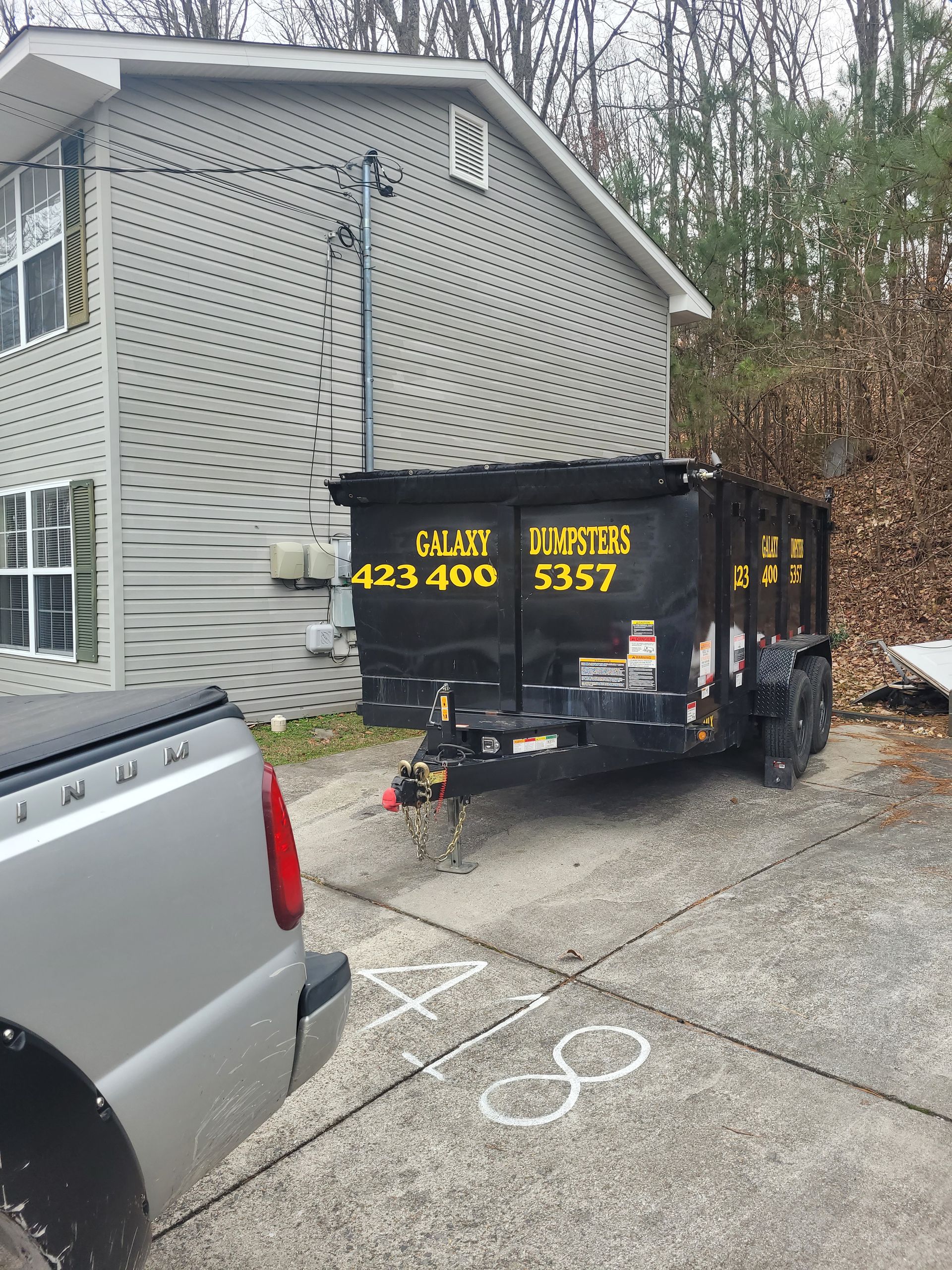A dumpster is parked in front of a house next to a truck.