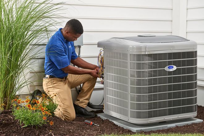 A service technician wearing a blue uniform kneels outside to repair a large, grey air conditioning unit.