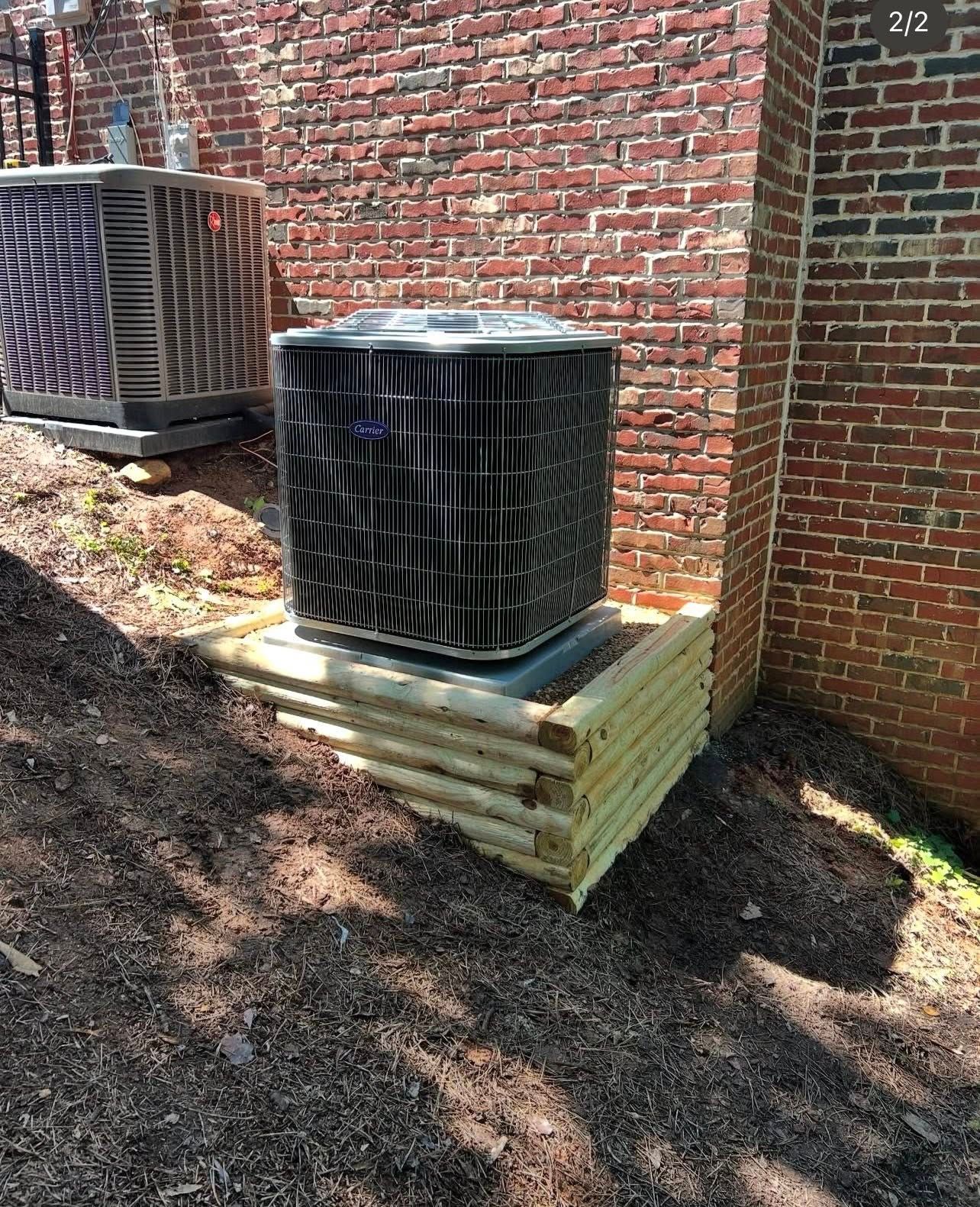 Two HVAC condenser units rest on wooden retaining walls against a brick exterior wall on a sloped, mulched landscape.
