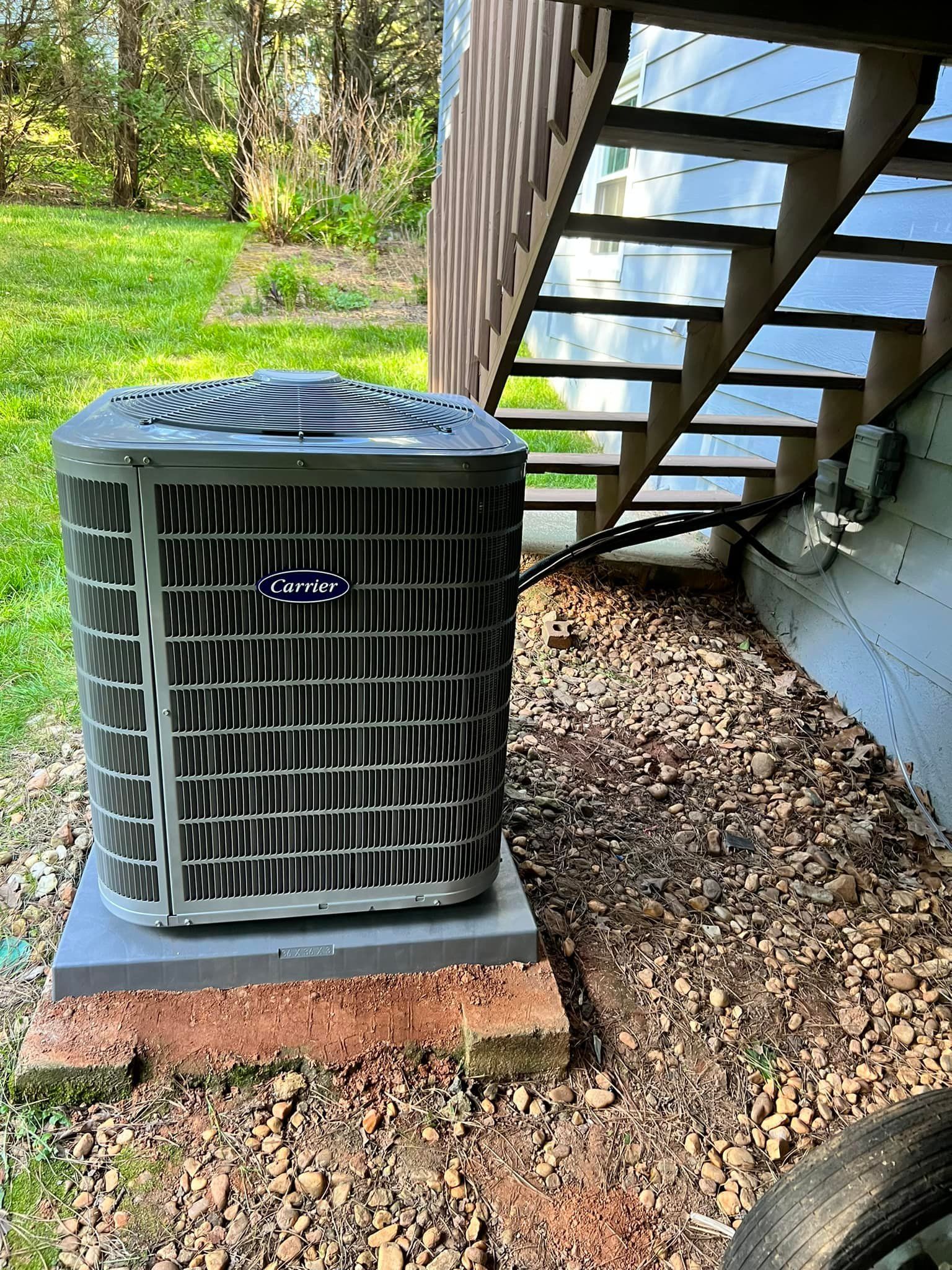 A gray Carrier outdoor air conditioning unit sits on a brick and concrete base beside a house with wooden stairs.