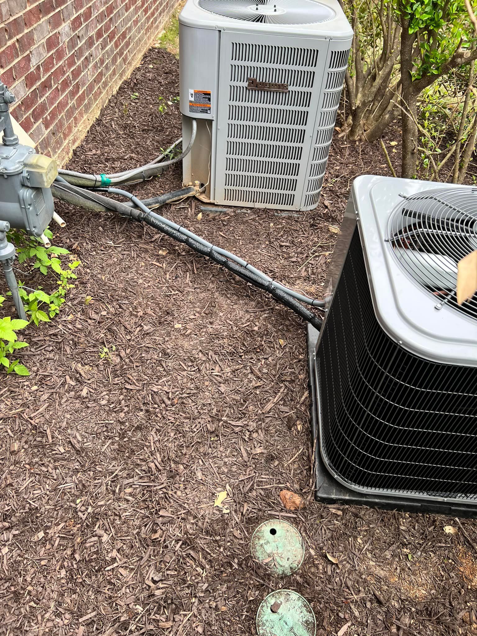 Two outdoor HVAC condenser units sit on a mulch-covered ground near a brick wall, connected by electrical conduits.