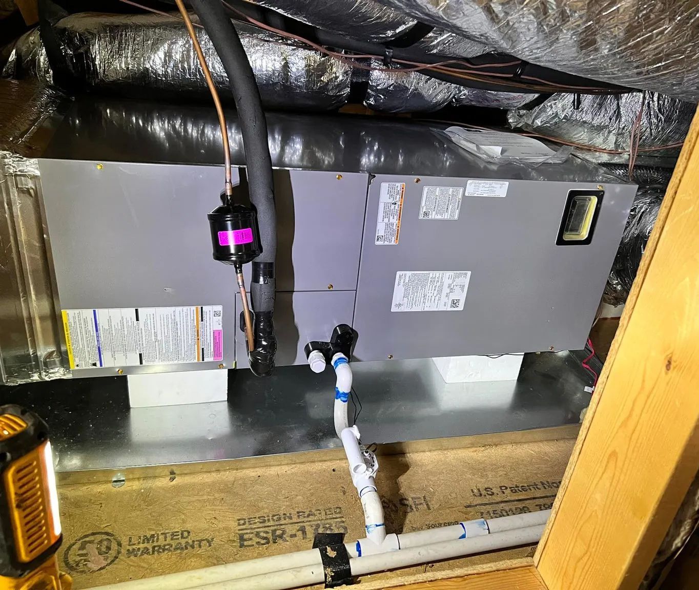 A gray HVAC air handler unit installed in an attic, featuring connected white PVC drain pipes and insulated refrigerant lines.