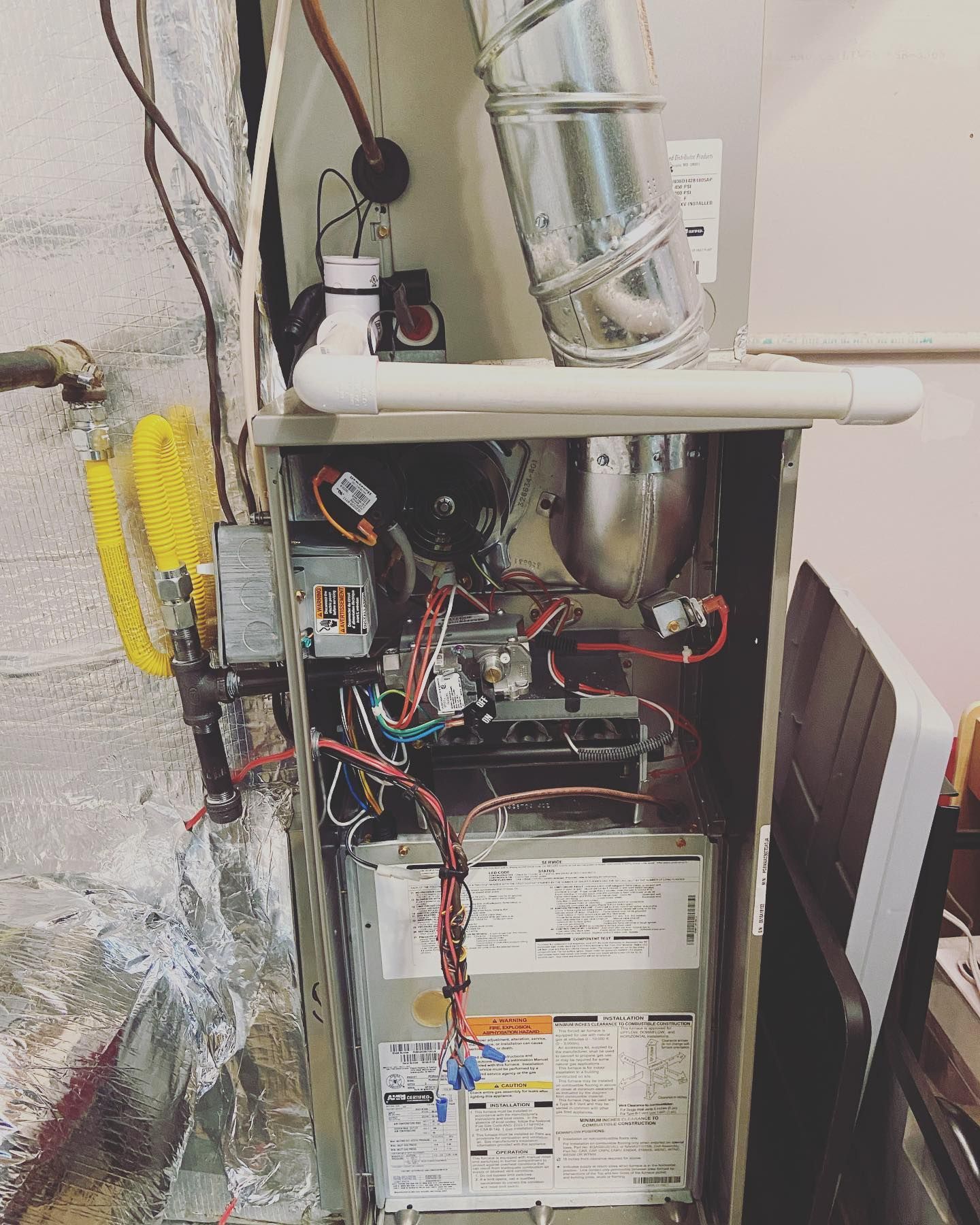 An open natural gas furnace in a utility space, showing internal wiring, a metal exhaust pipe, and a control panel label.