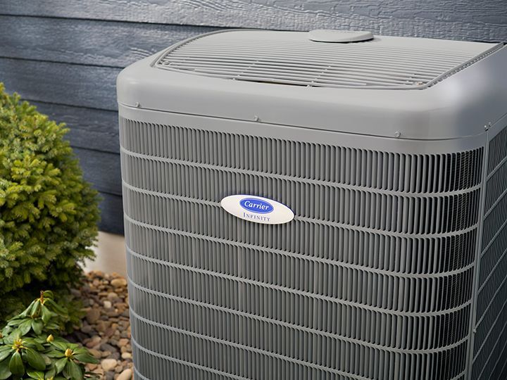 A grey Carrier central air conditioning unit positioned outside against a blue-sided house near a green shrub.