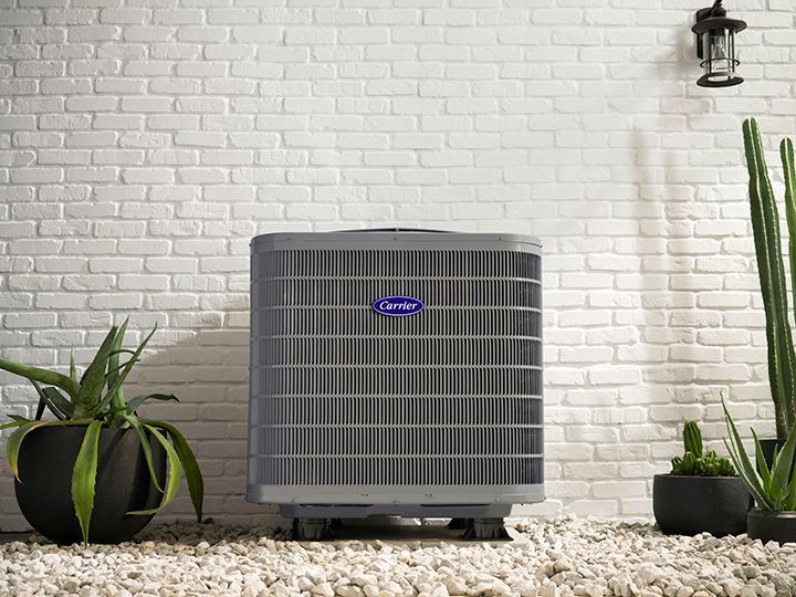 A grey Carrier air conditioning unit sits on a gravel floor against a white brick wall, flanked by potted plants.