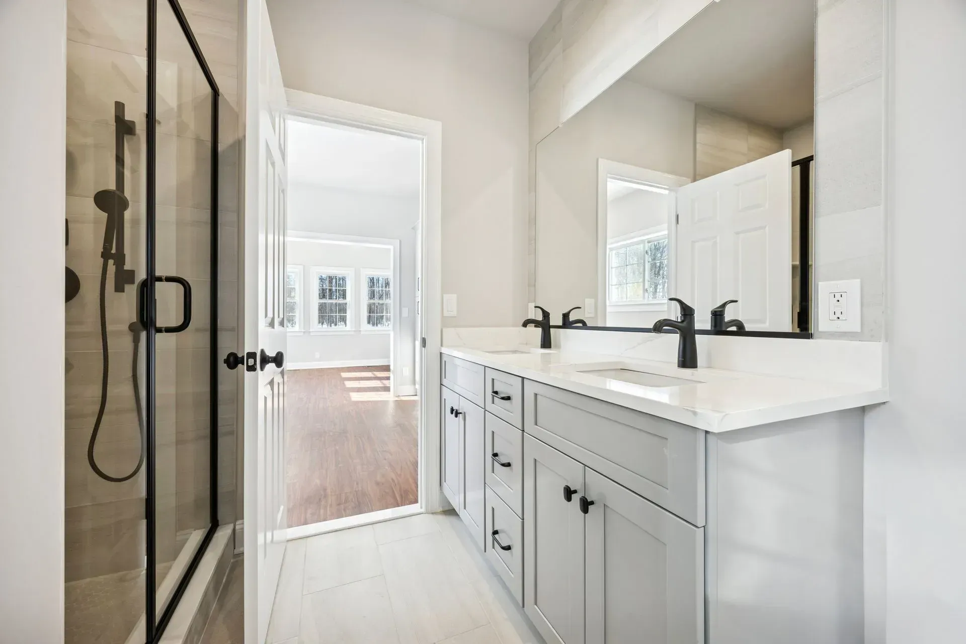 Modern bathroom with glass shower, double vanity, and open door to a bright bedroom