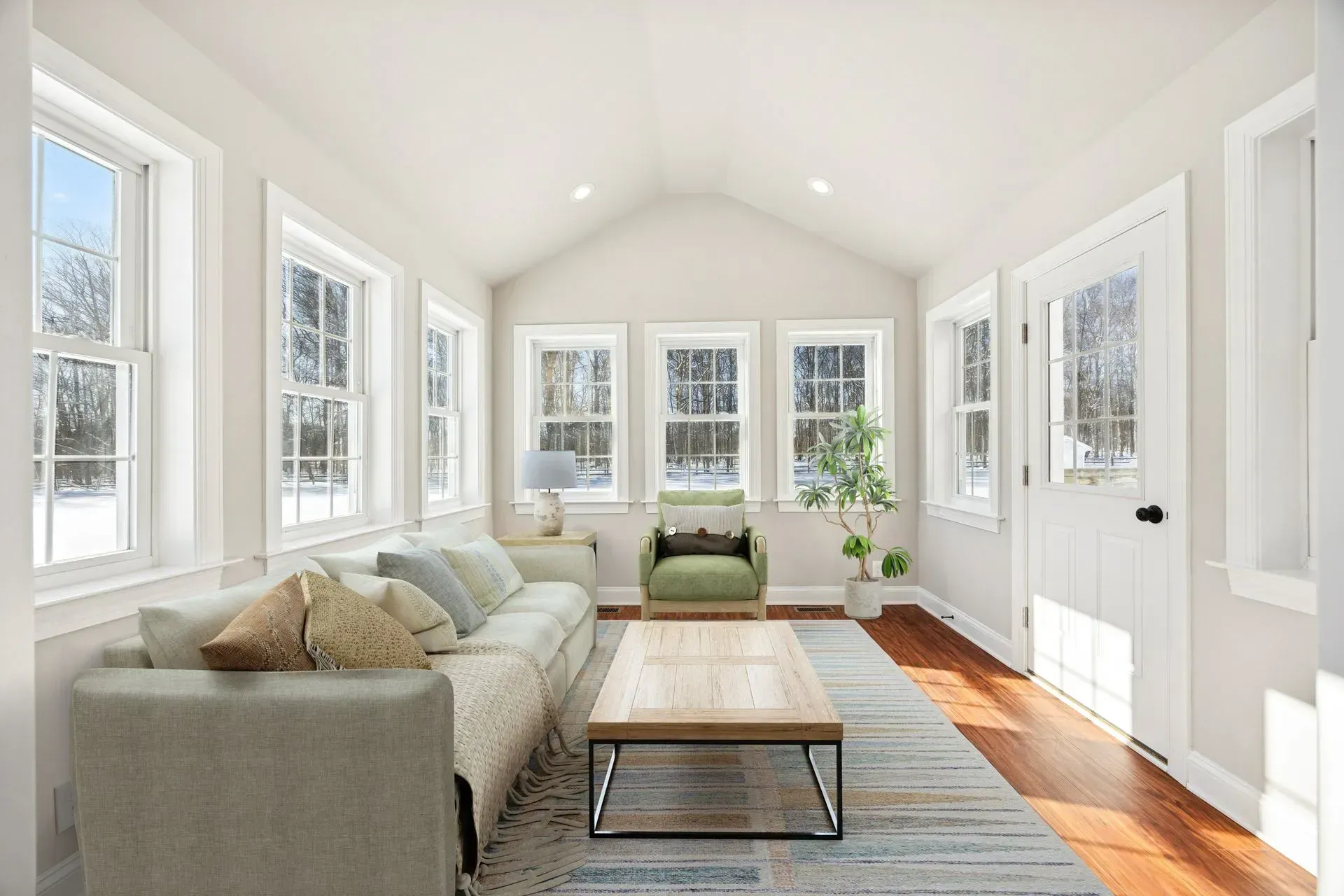 Bright sunroom with white walls, large windows, a gray sofa, armchair, coffee table, and wood floor.