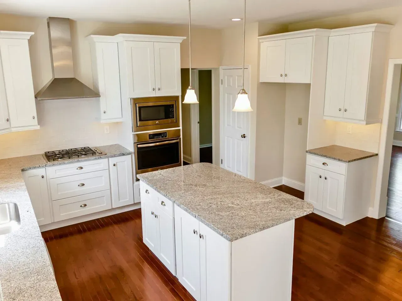 Bright white kitchen with granite island, white cabinets, stainless oven, and warm wood floors