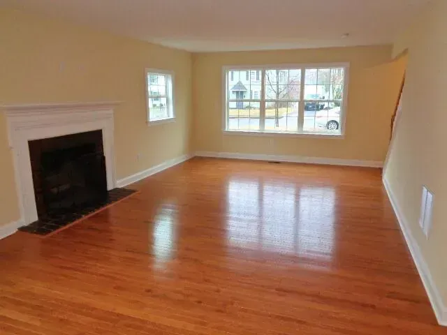 Empty living room with hardwood floors, beige walls, fireplace, and large front window.
