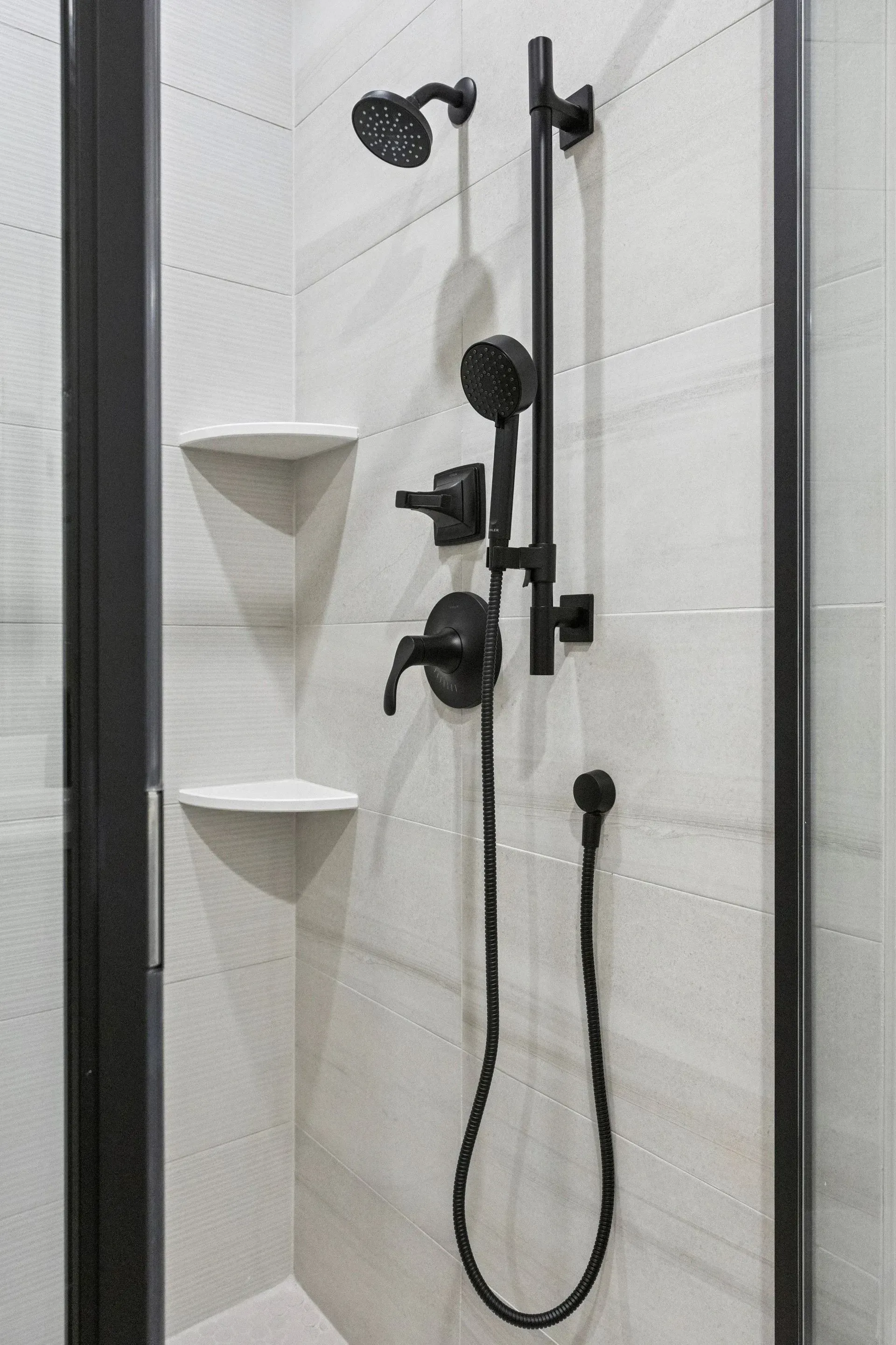 Bathroom shower with black grab bars, handheld showerhead, and white corner shelves.