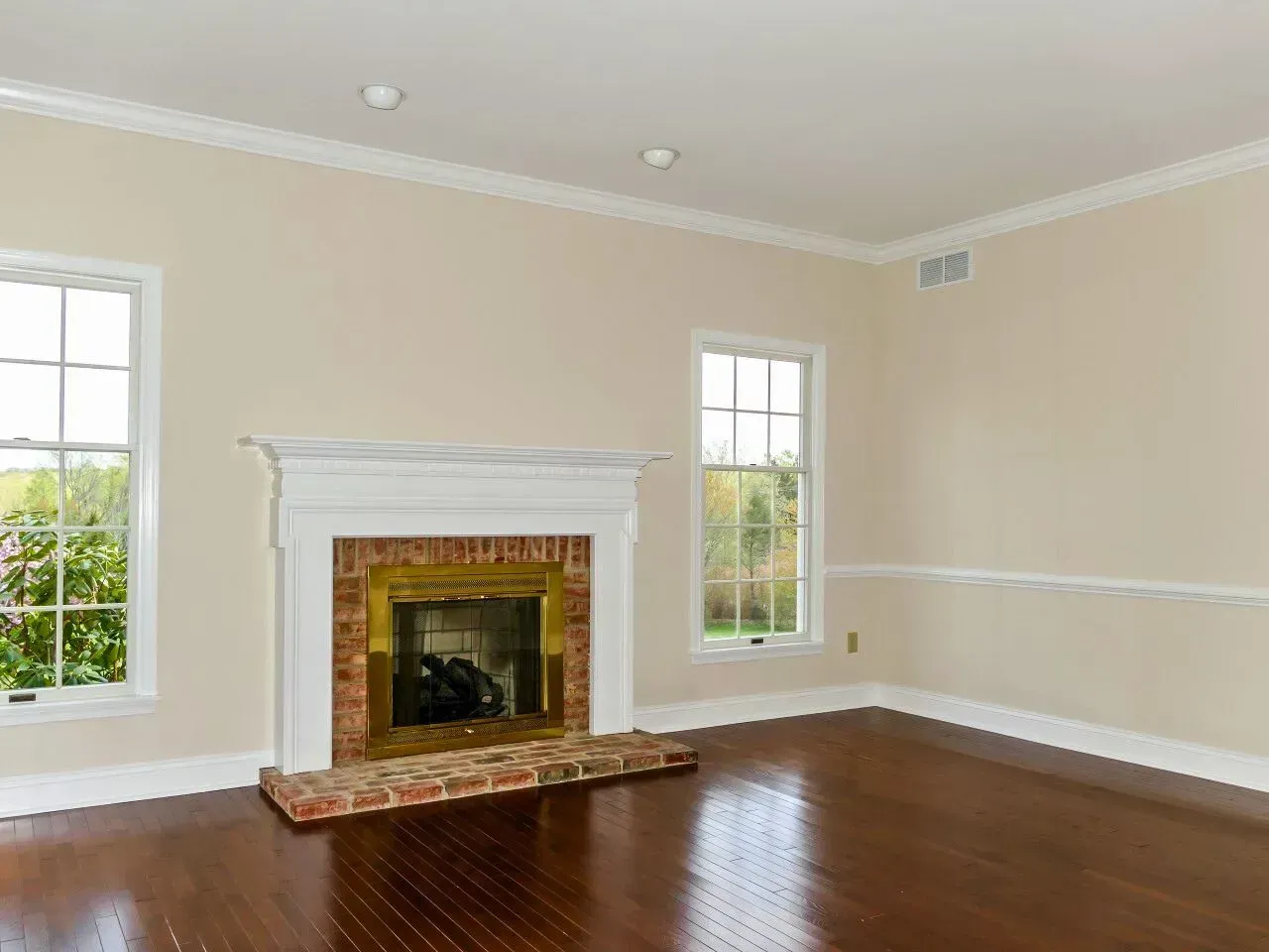 Empty beige living room with hardwood floors, white trim, and a brick fireplace between two windows