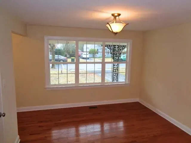 Empty beige room with hardwood floor, ceiling light, and large window overlooking a yard.