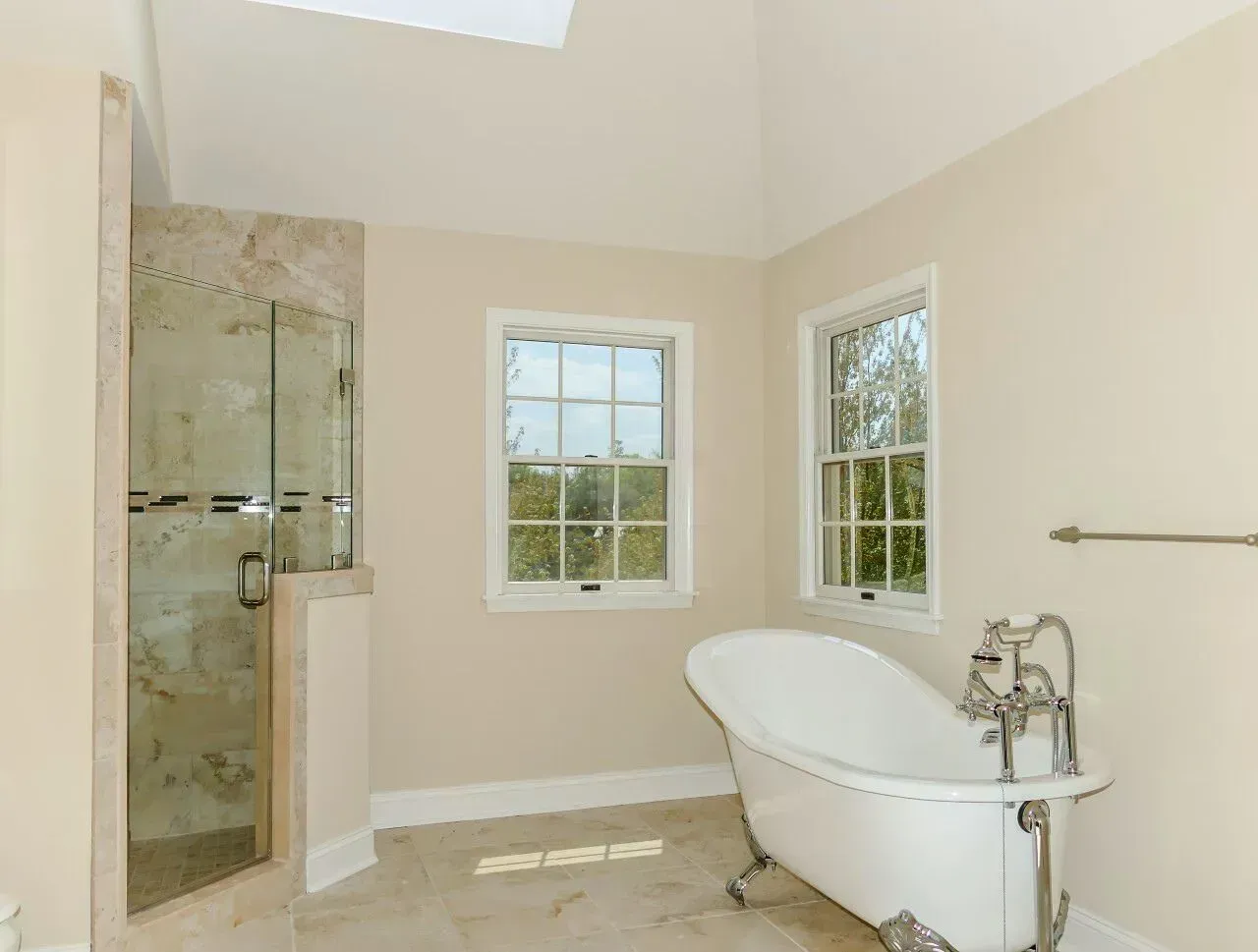 Bright bathroom with a glass shower, freestanding tub, beige walls, and two windows