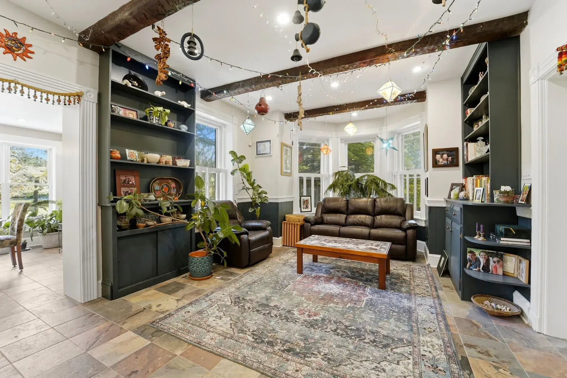 Bright living room with a brown sofa, patterned rug, plants, shelves, and hanging decor near large windows