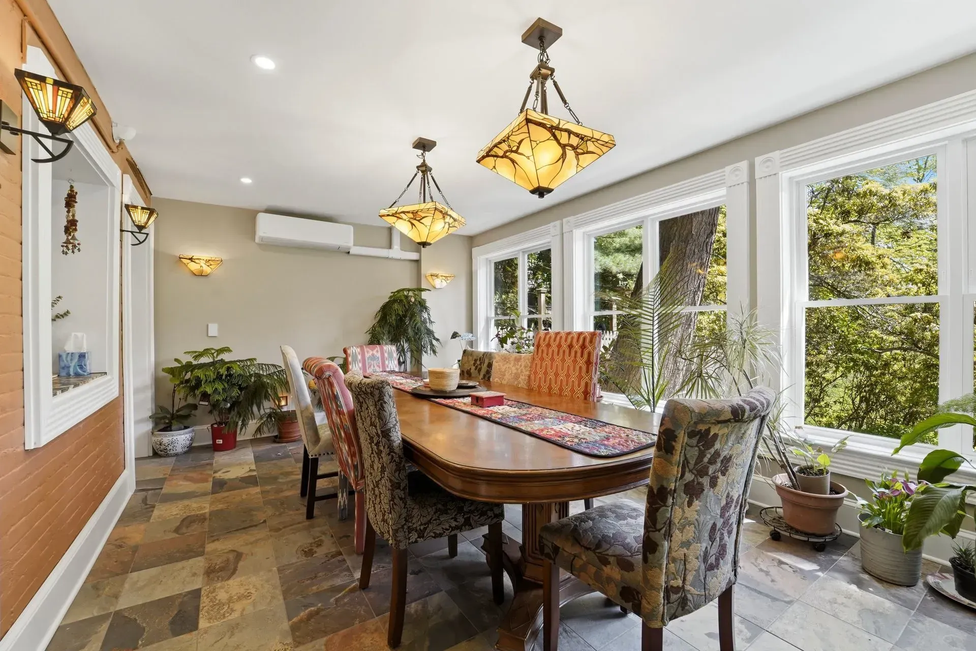 Bright dining room with a wooden table, patterned chairs, hanging lamps, and large windows overlooking greenery
