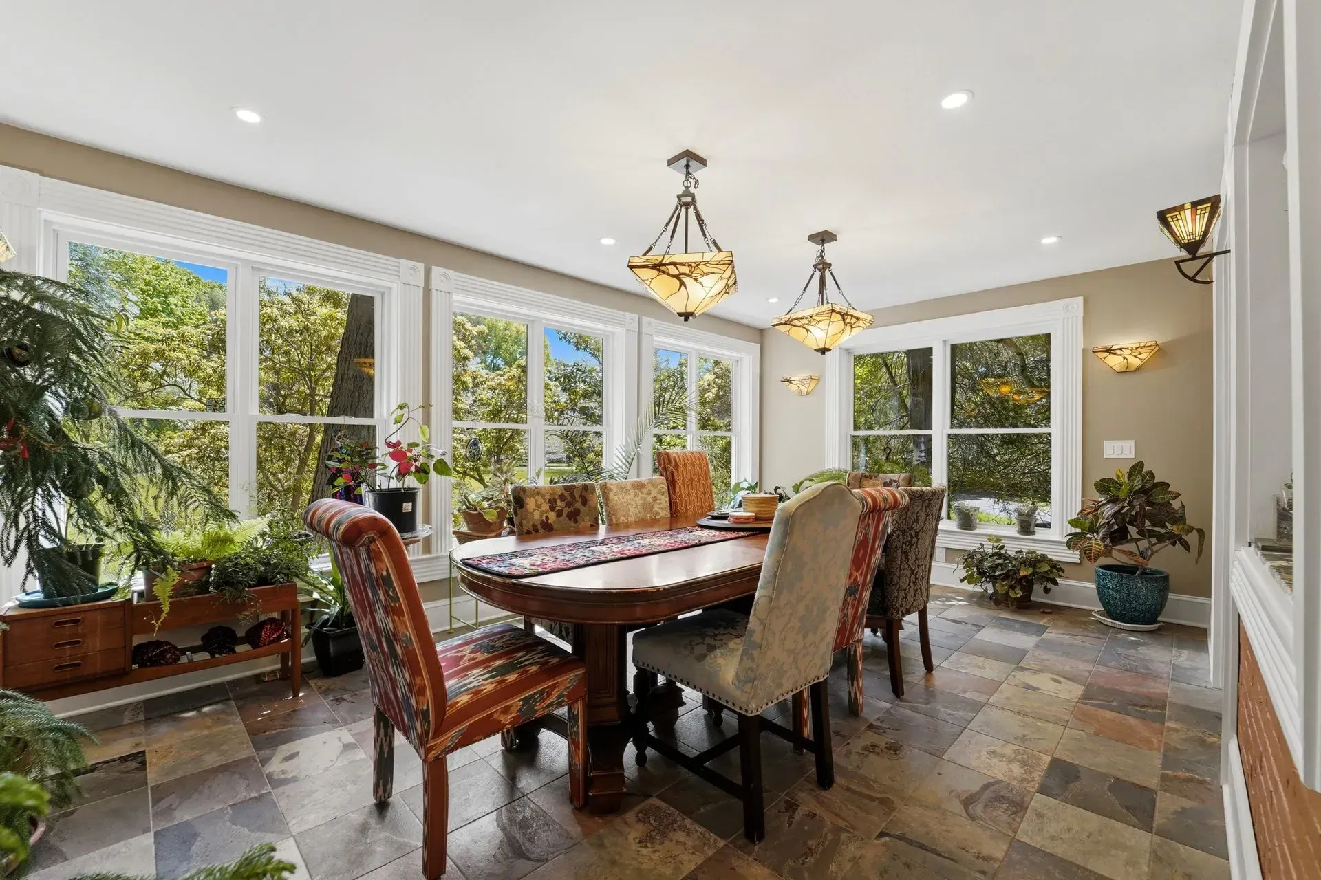 Bright dining room with a long table, patterned chairs, chandeliers, and large windows overlooking greenery