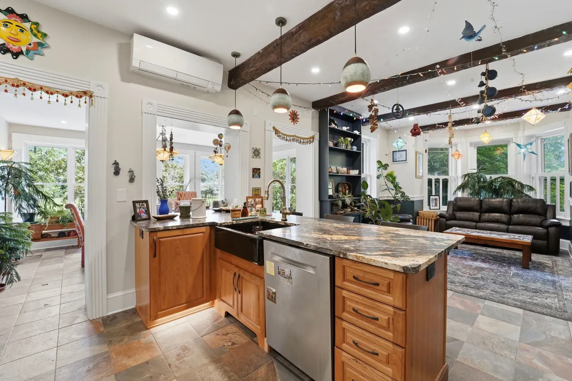 Bright, open kitchen with island and stainless dishwasher, facing a cozy living room with wood beams and dark sofa