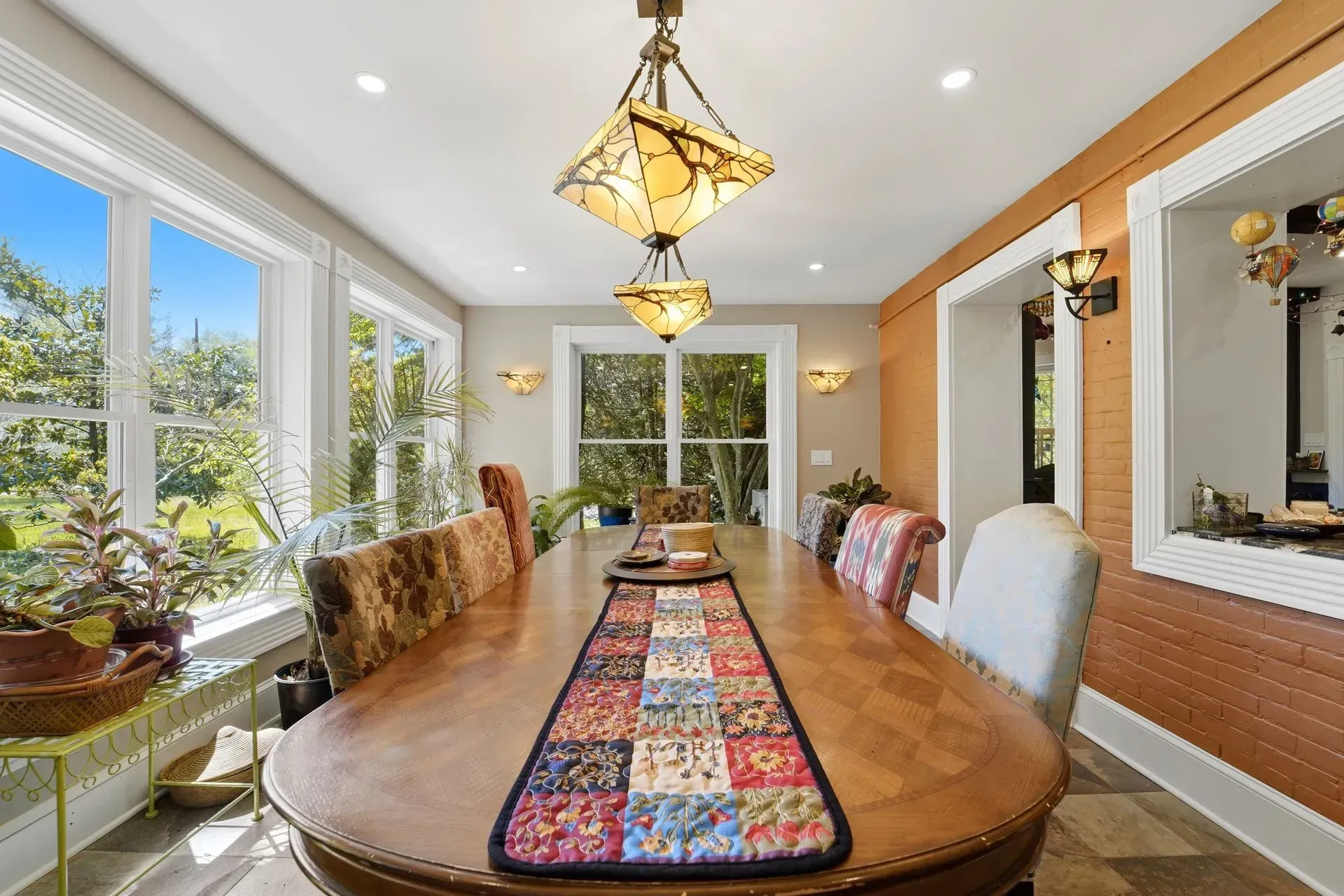 Bright dining room with long wooden table, colorful runner, chandelier, and large windows overlooking greenery