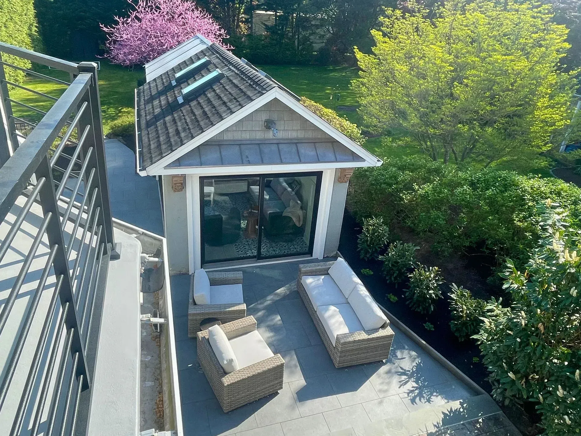 Modern patio by a small white cottage, with wicker seating and surrounding green shrubs and pink blossoms
