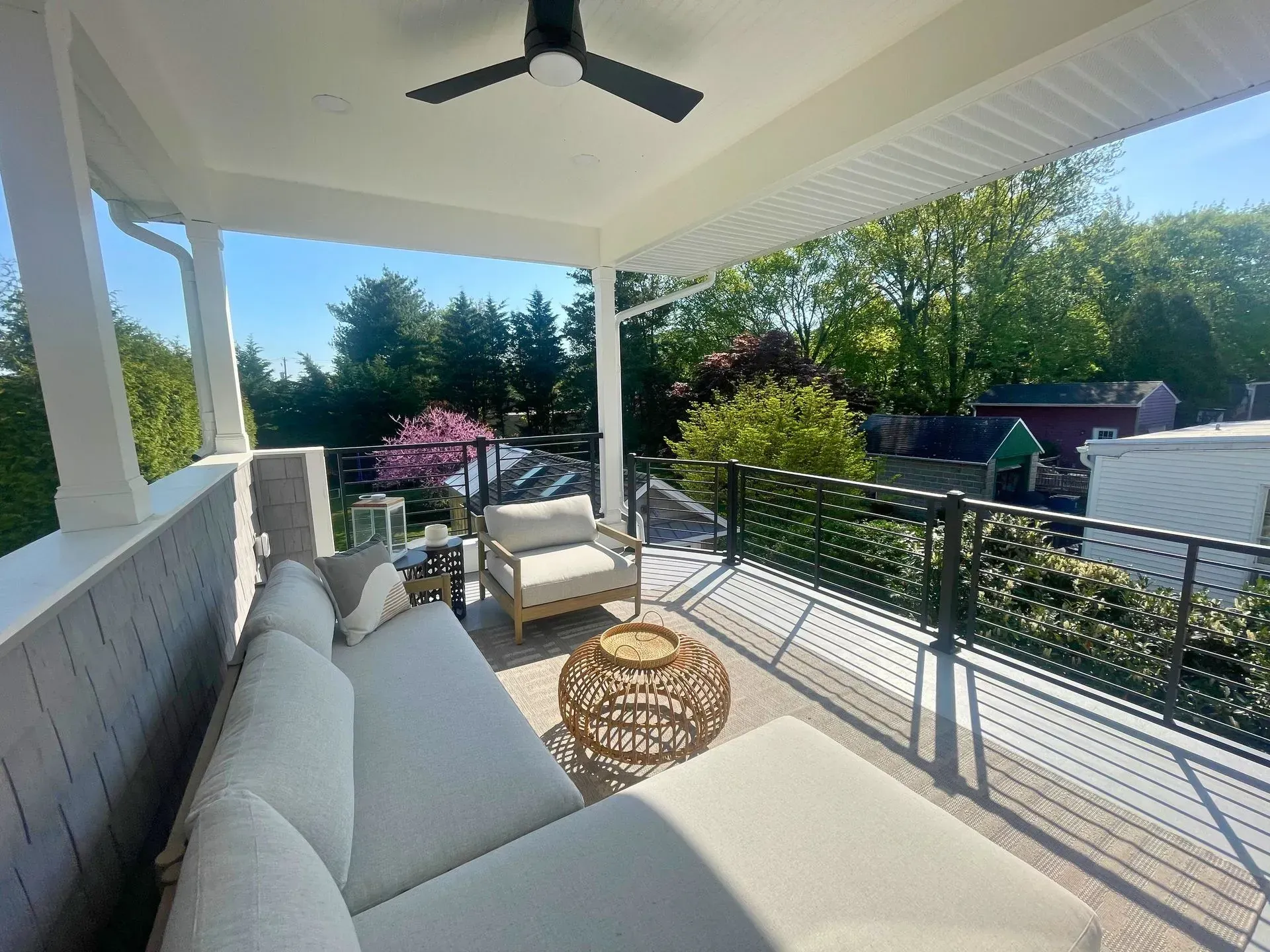 Covered patio with white outdoor seating, wicker table, and a view of green trees and neighboring houses.