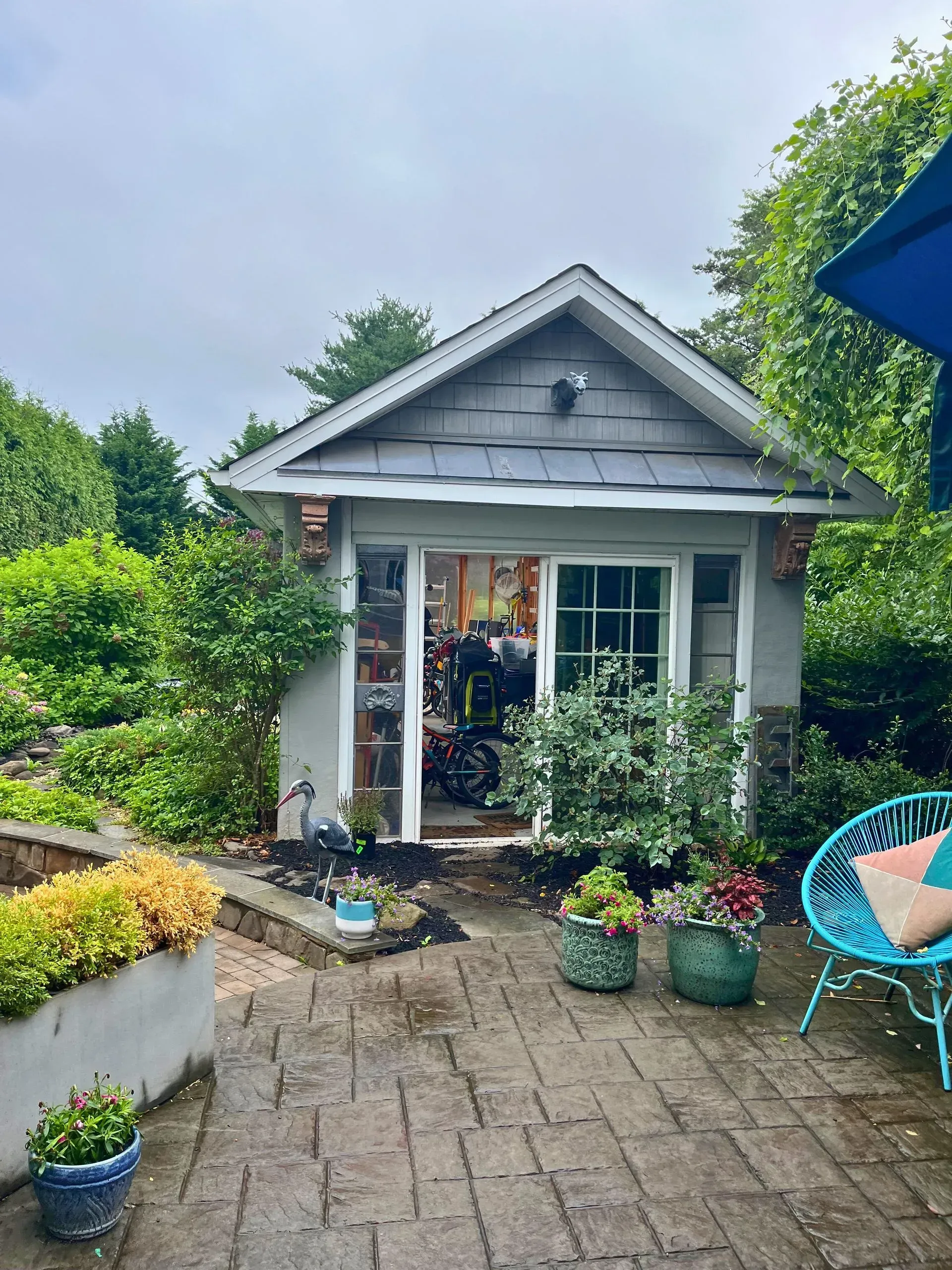 Small garden shed with open door, surrounded by potted plants and a brick patio.
