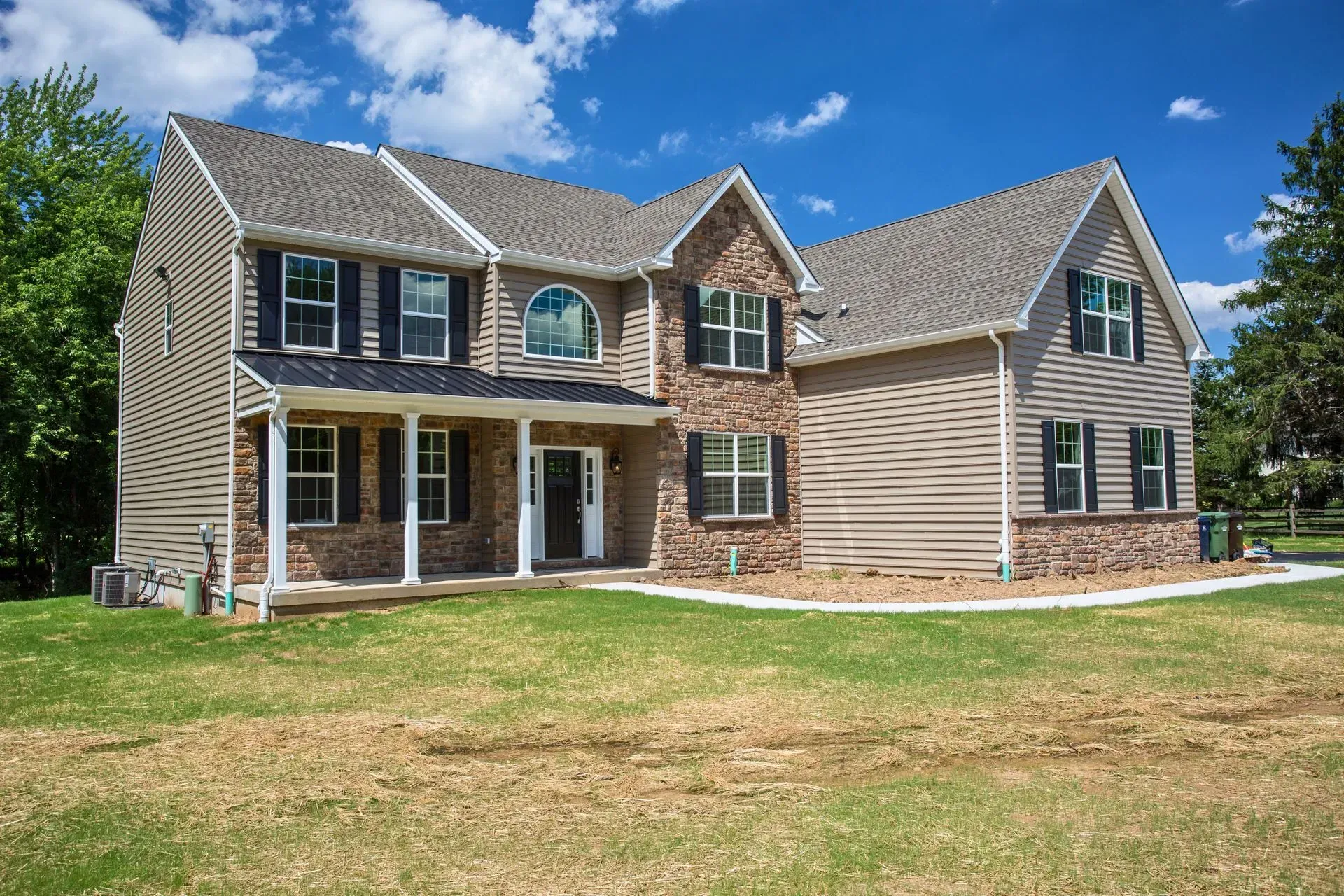 Two-story tan house with brown shutters and a large front lawn under a blue sky
