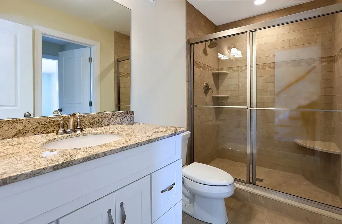 Modern bathroom with granite vanity, mirror, toilet, and glass-enclosed shower on beige tile floor