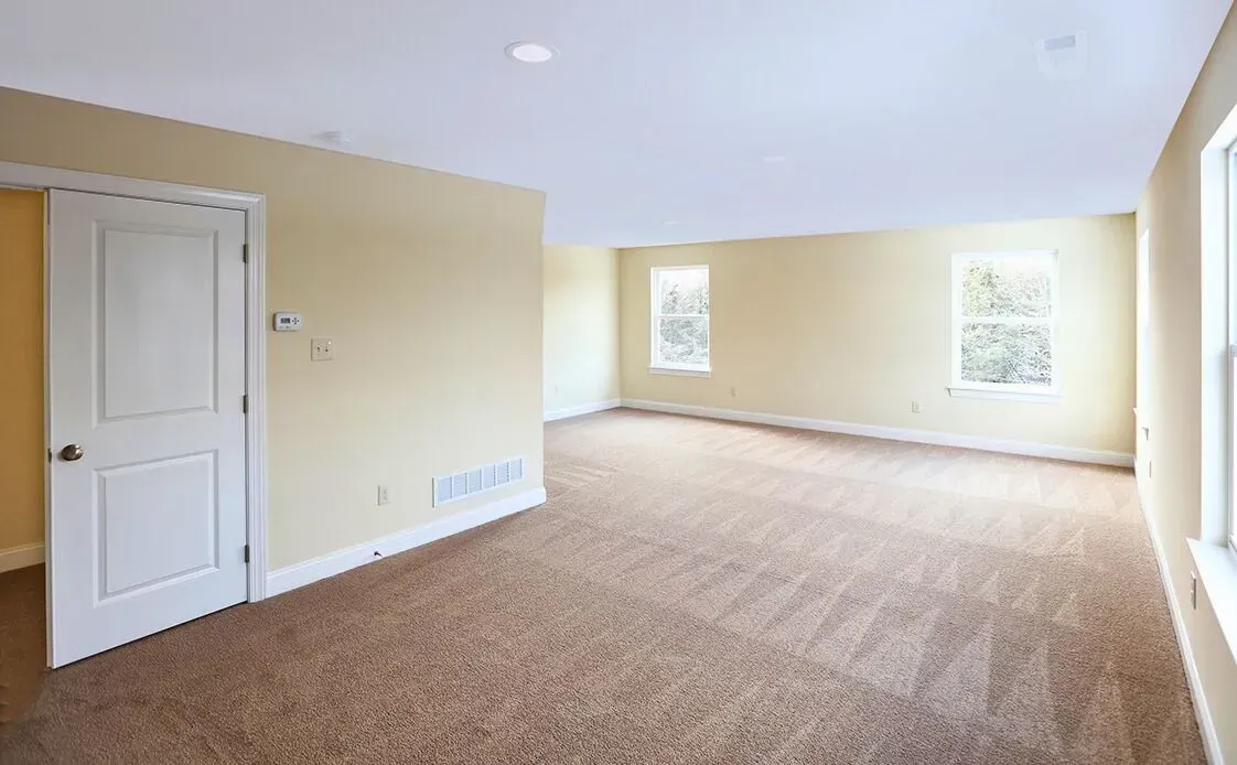 Empty beige living room with carpet, white door, and two windows letting in natural light