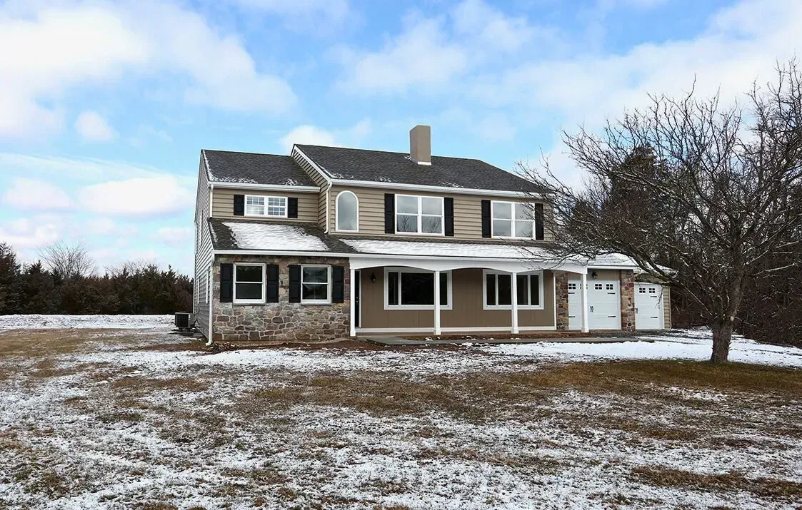 Snow-covered two-story farmhouse with a front porch and leafless tree in a winter field