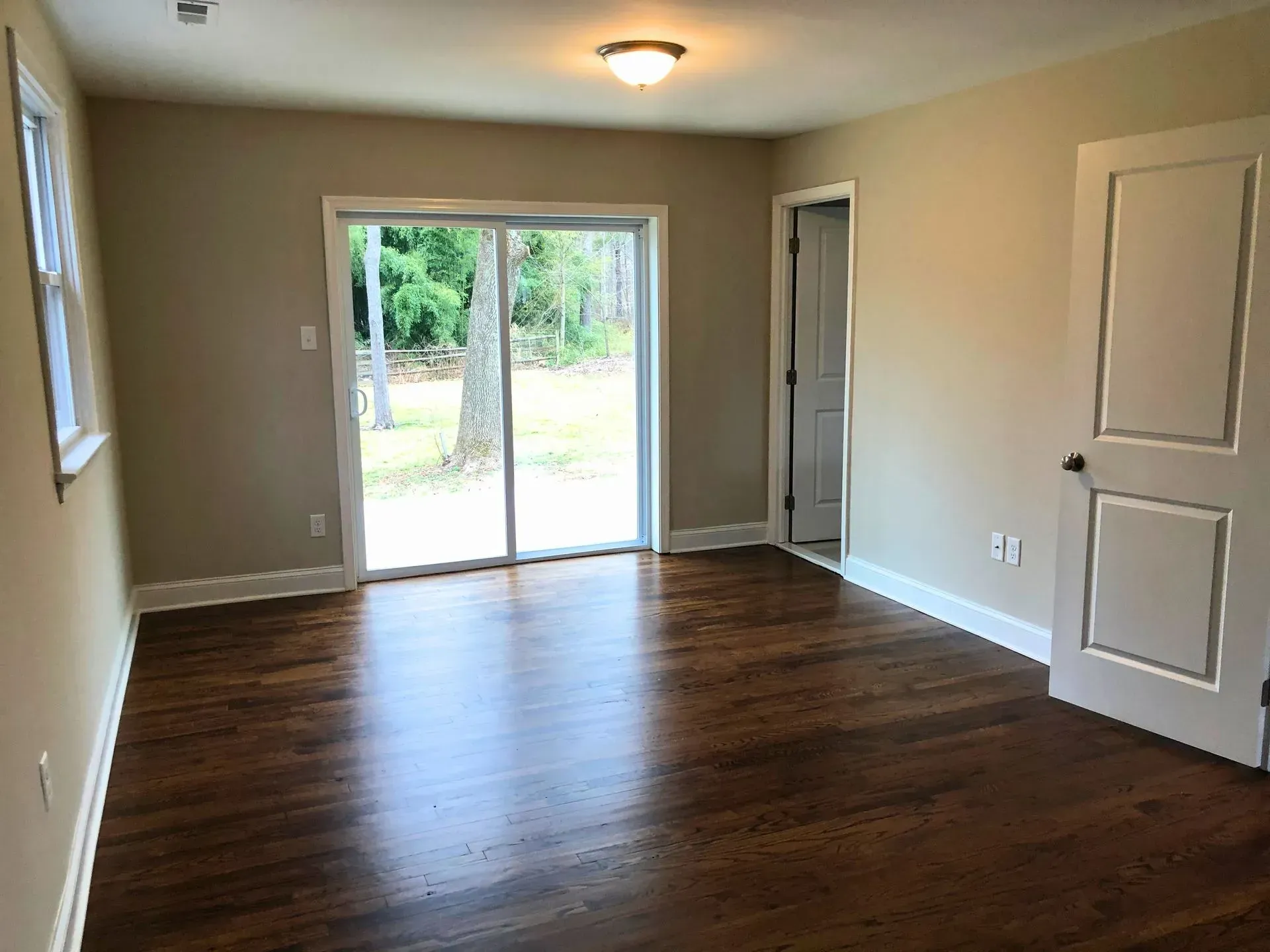 Empty beige room with dark wood floors and sliding glass doors opening to a backyard.