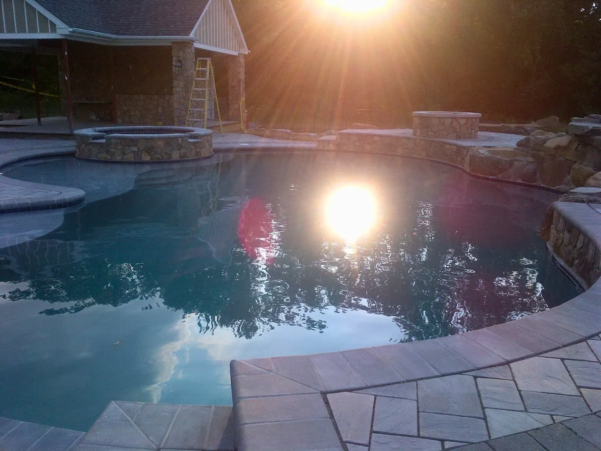 Backyard pool at sunset with bright sun reflecting on the water and stone patio edge