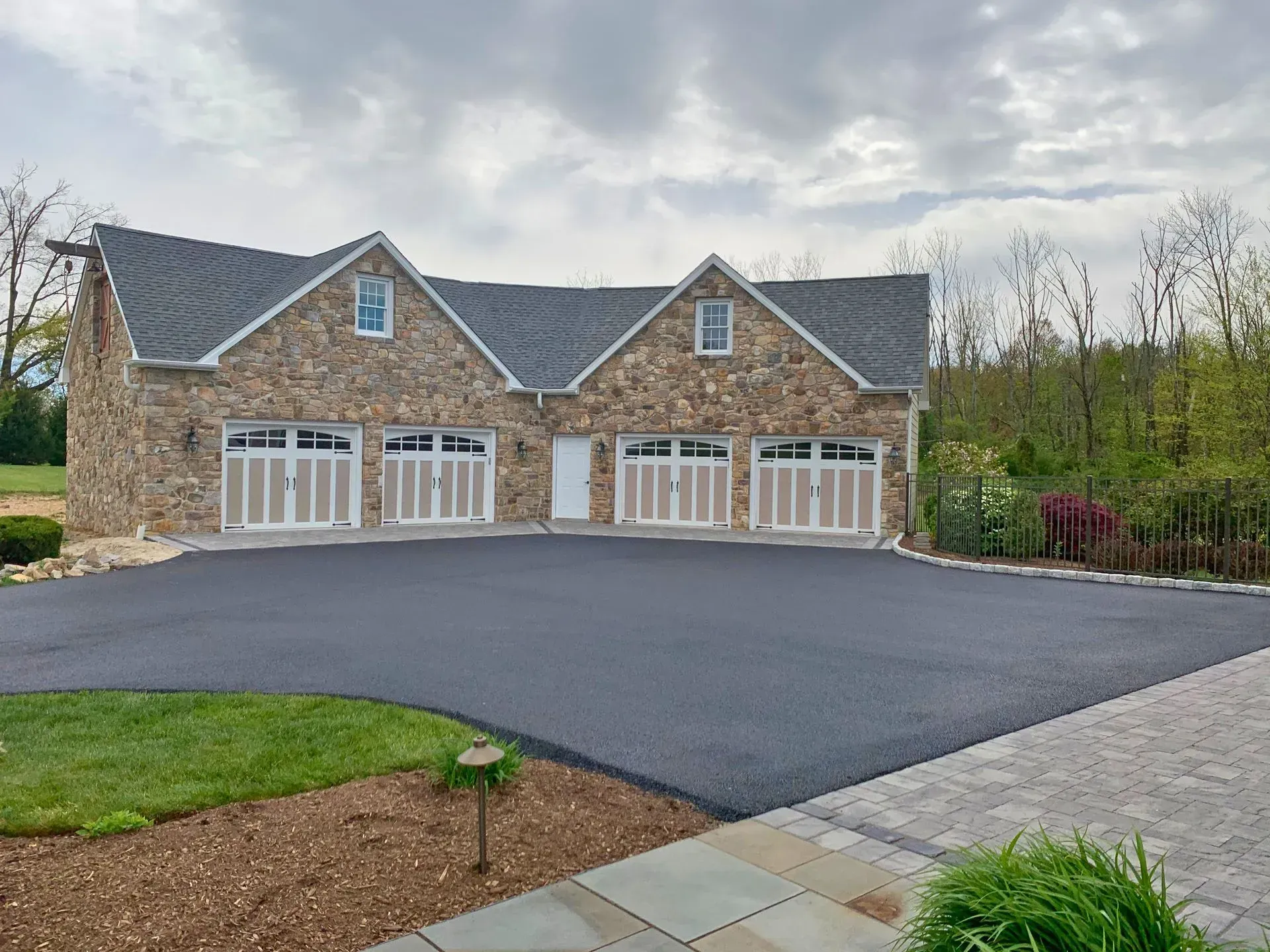 Large stone house with multiple garage doors, dark roof, and a broad paved driveway under a cloudy sky