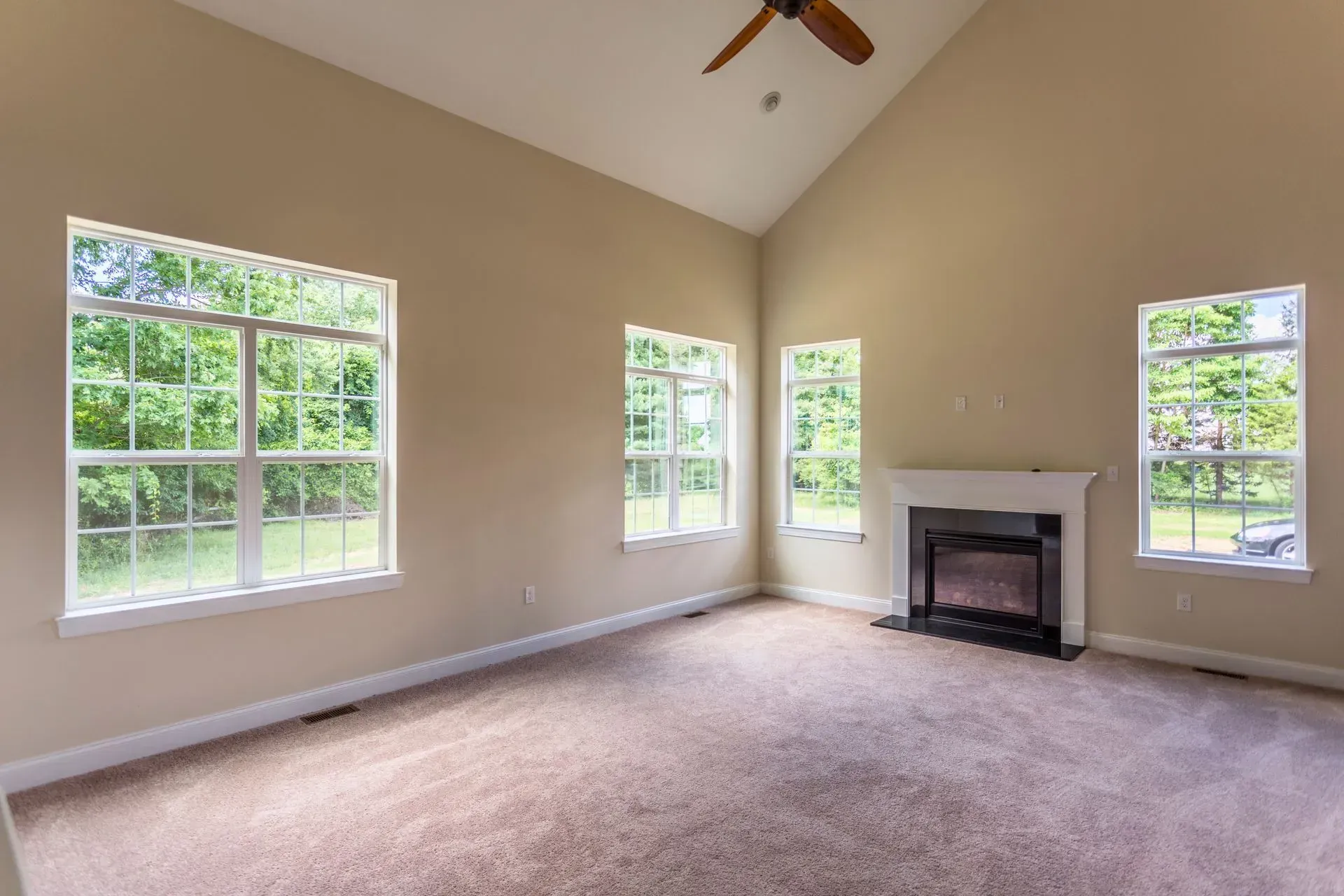 Empty beige living room with carpet, large windows, and a fireplace under a ceiling fan
