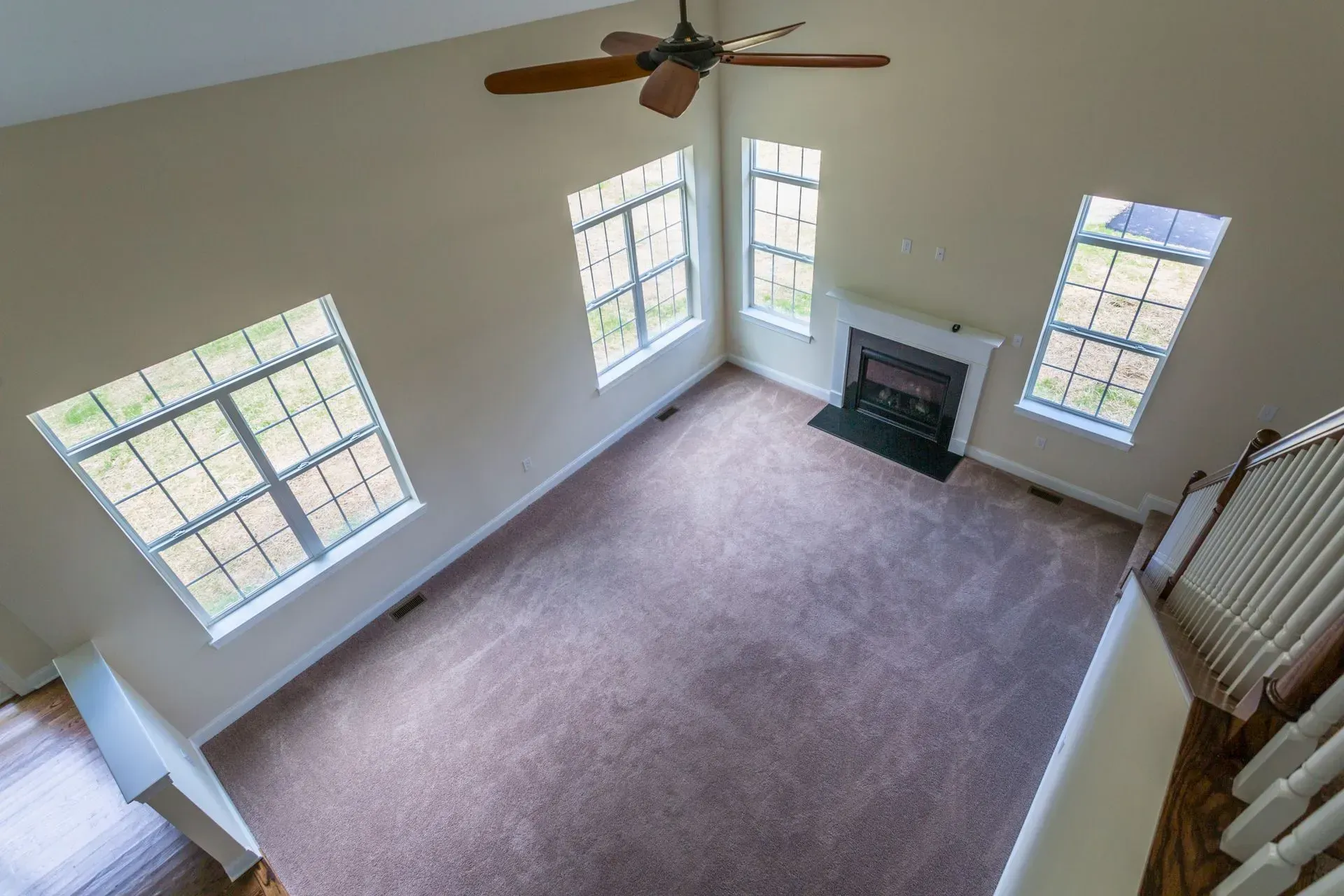 Empty beige living room with carpet, large windows, ceiling fan, and fireplace.