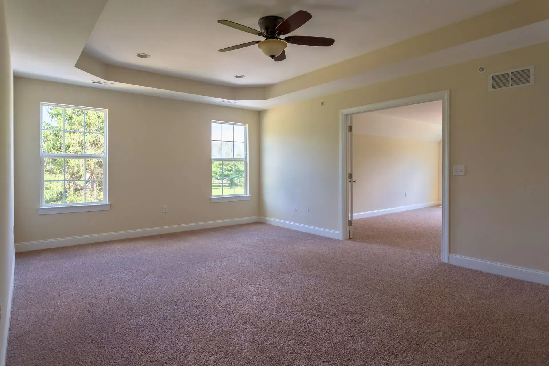 Empty beige carpeted living room with ceiling fan, two windows, and an open doorway to another room
