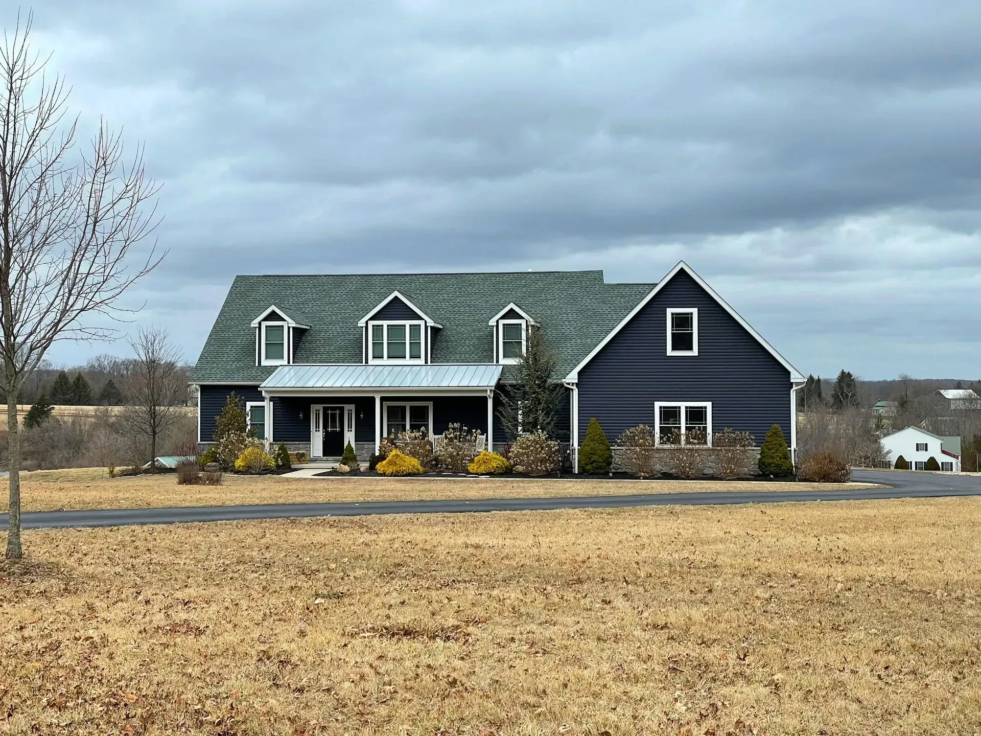 Dark blue farmhouse with white porch sits in a field under cloudy skies.