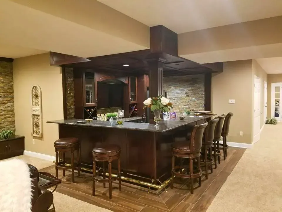 Spacious home bar with dark wood cabinetry, bar stools, granite counter, and beige carpeting in an open interior.