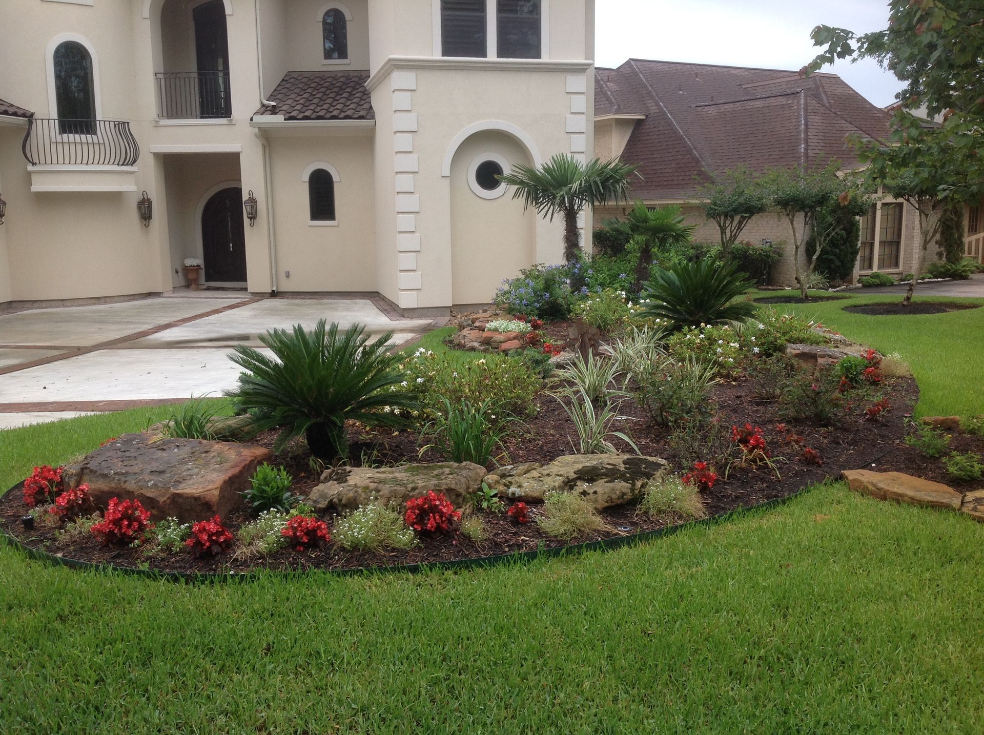 A landscaped front yard with a variety of plants and red flowers in front of a beige house.