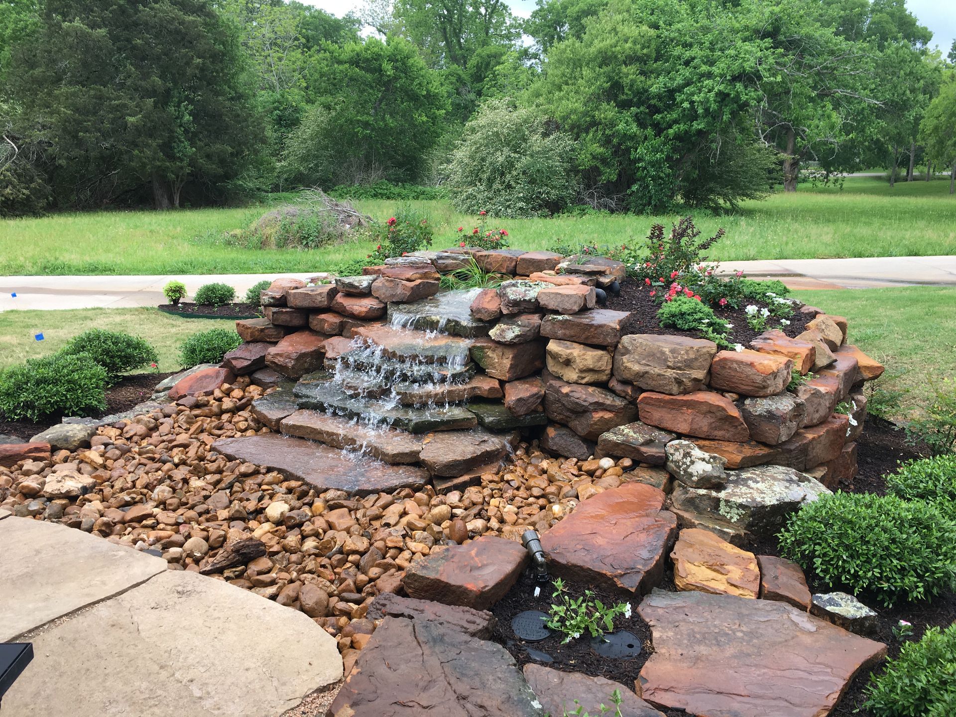 Stone waterfall feature with cascading water, surrounded by rocks, plants, and green grass.