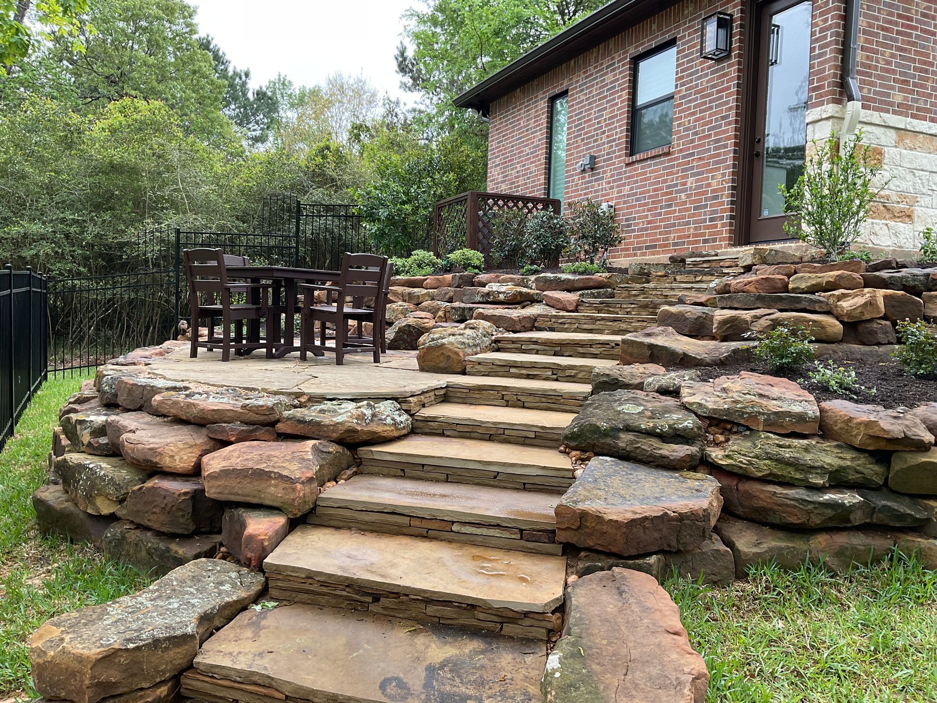 Stone steps leading up to a patio with a table and chairs next to a brick house and green foliage.