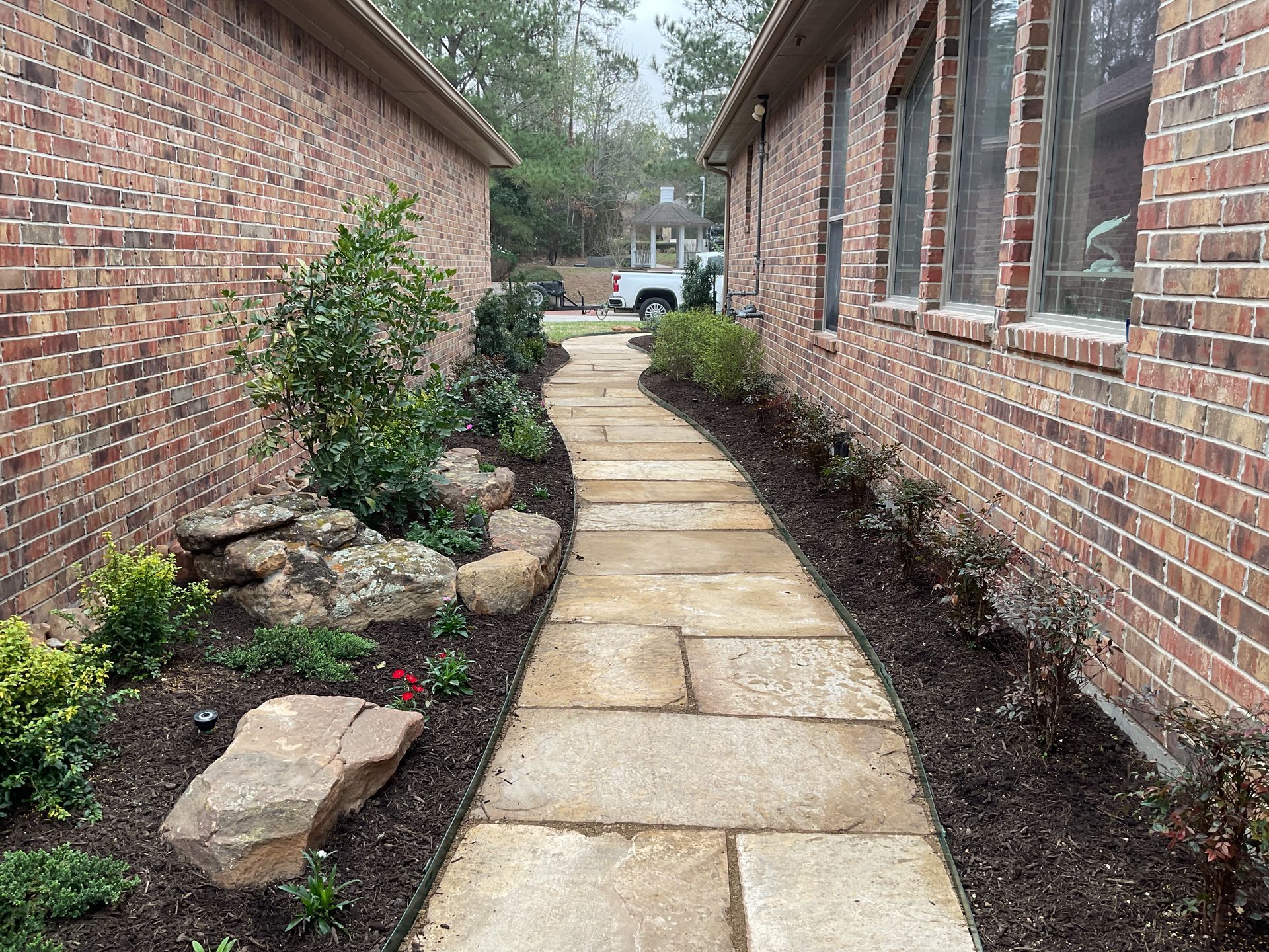 Stone pathway between two brick buildings, lined with plants and mulch.