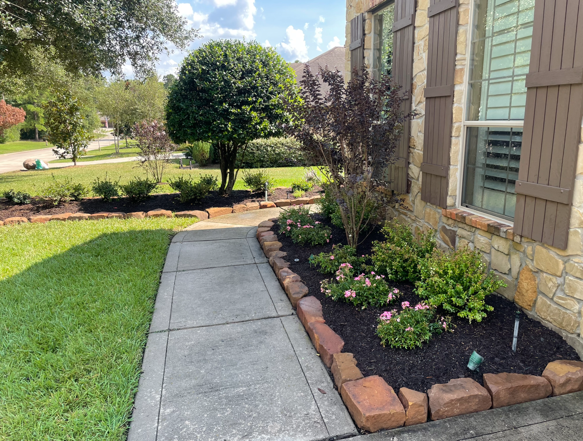 A brick-lined flower bed with dark mulch and pink flowers next to a sidewalk and house.