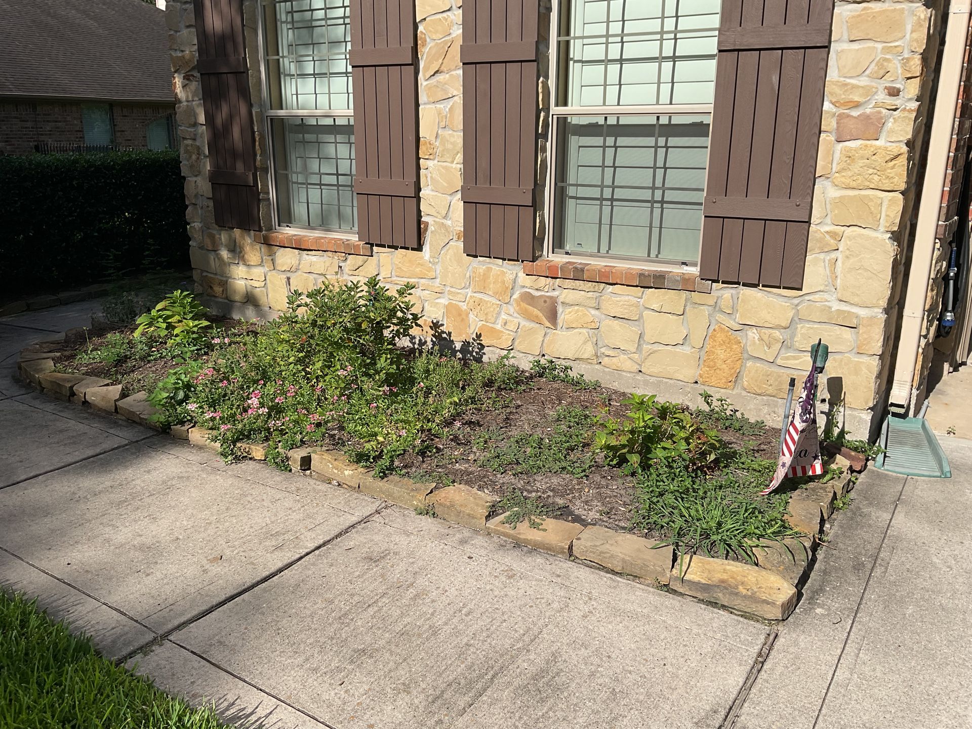 Flower bed in front of a stone house, with brown shutters and American flag.