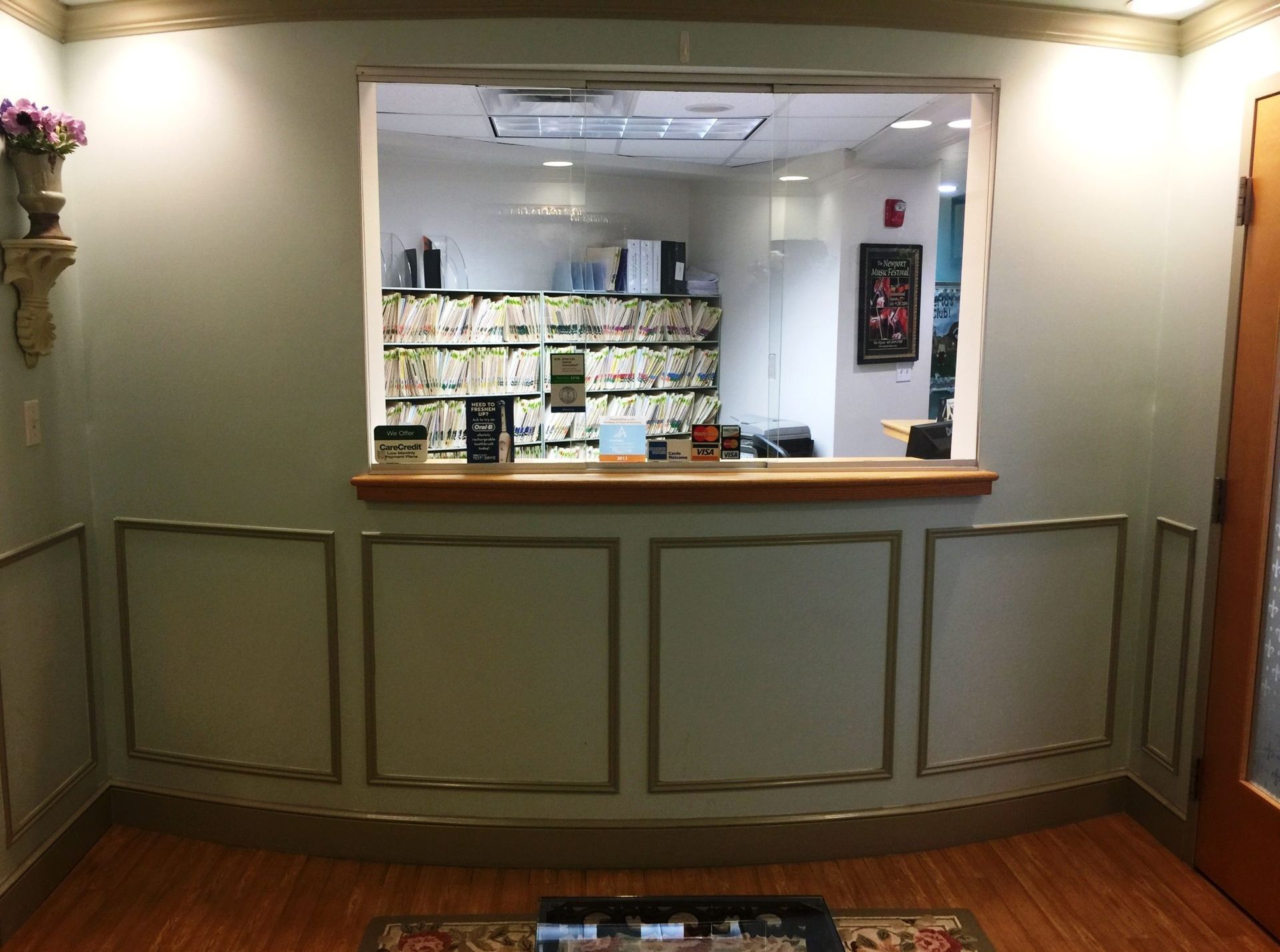 Reception area with a window to a work area and a door, pale green walls, and wood-look flooring.