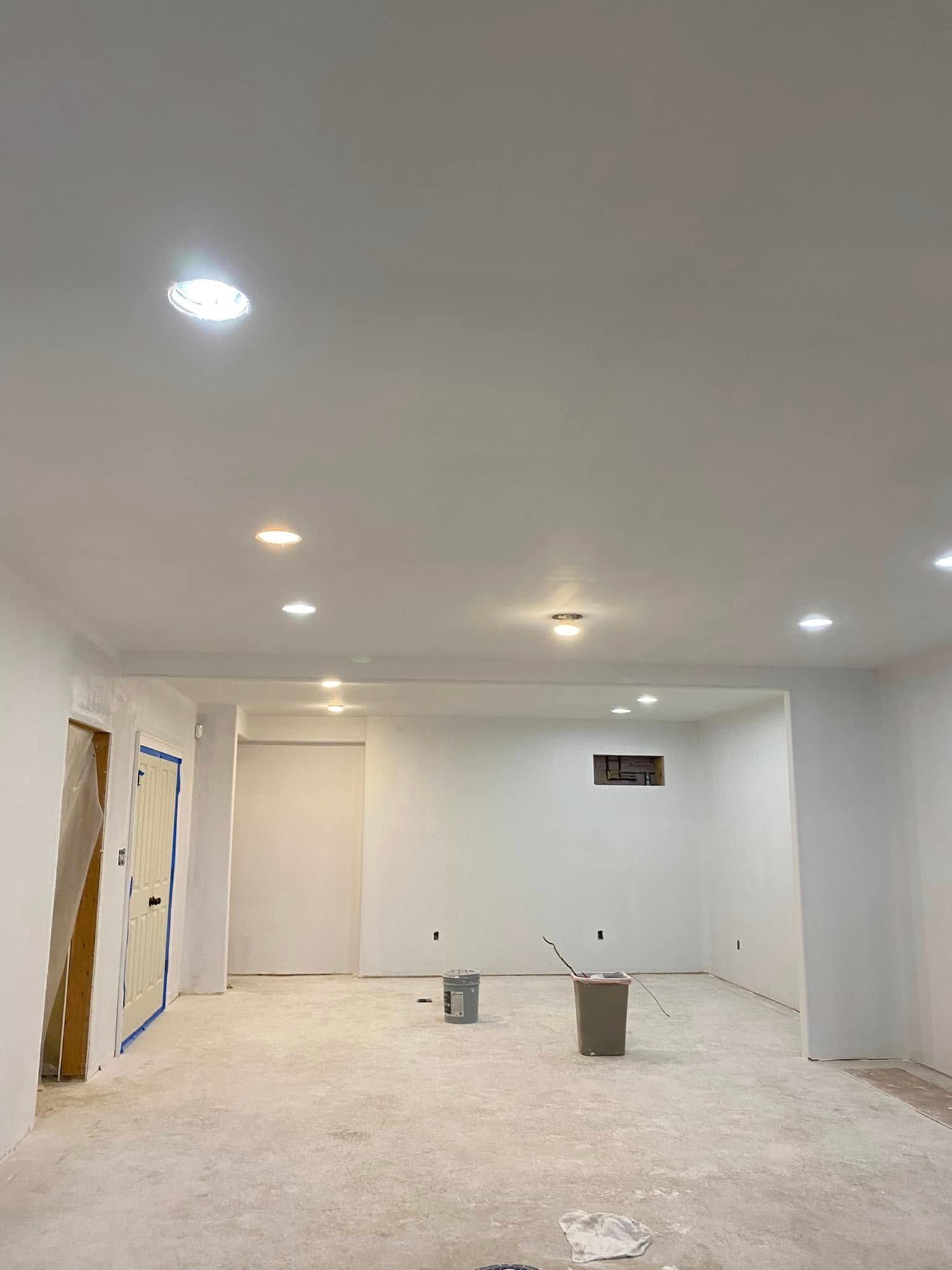 White-walled room with recessed lighting. Unfinished floor, doors, and construction materials are present.