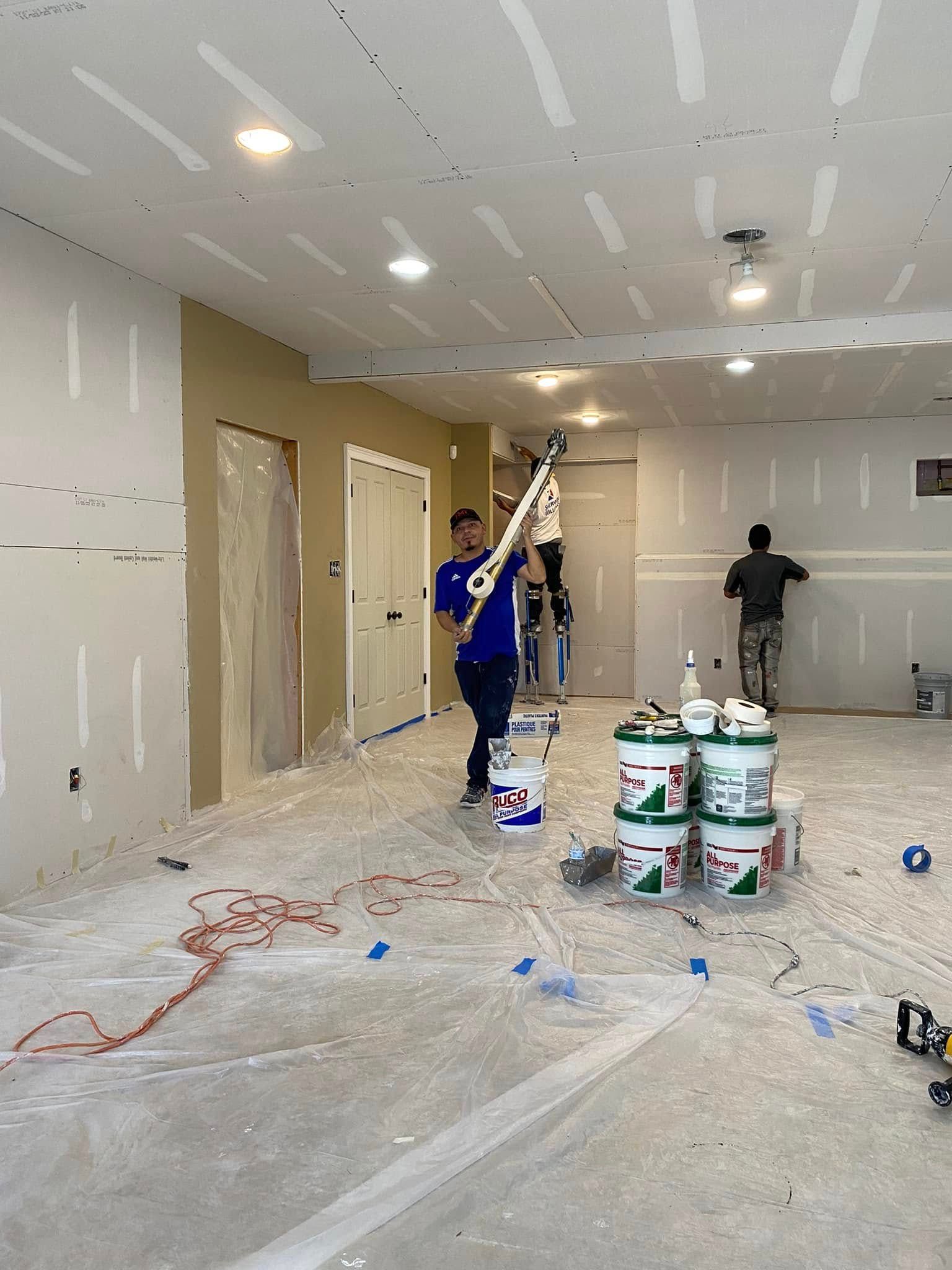 Two men drywalling a room under construction; white walls and ceiling; paint buckets.