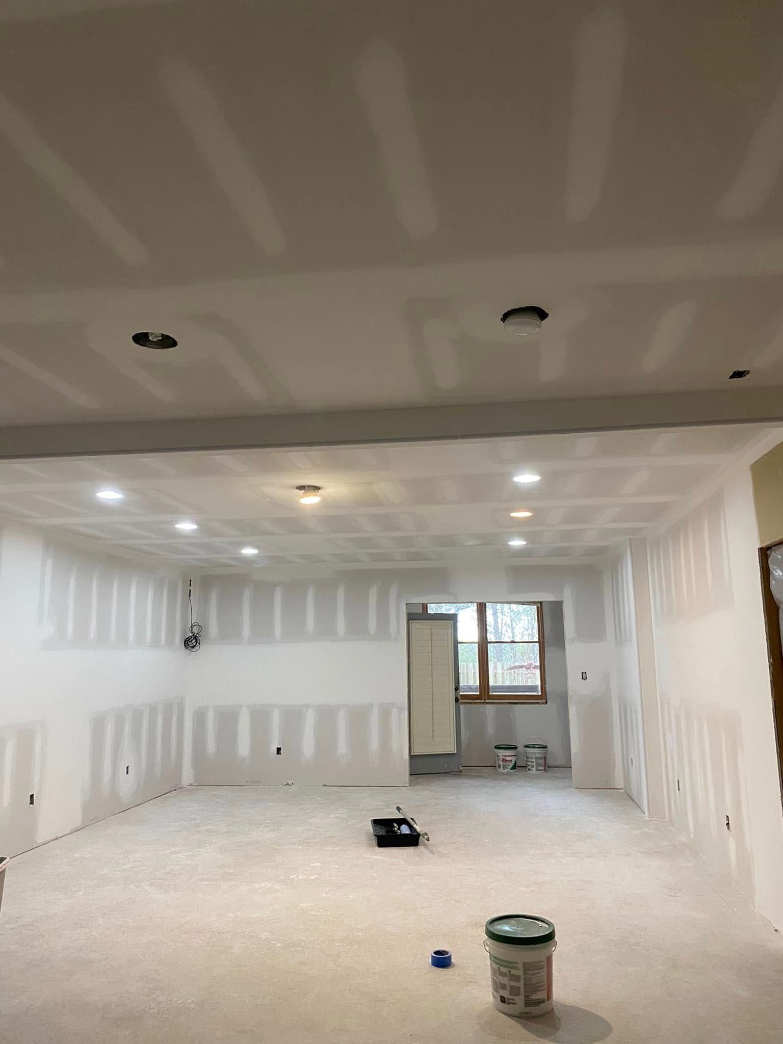 Interior room under construction with drywall on walls and ceiling. Recessed lights, paint can, and tools visible.
