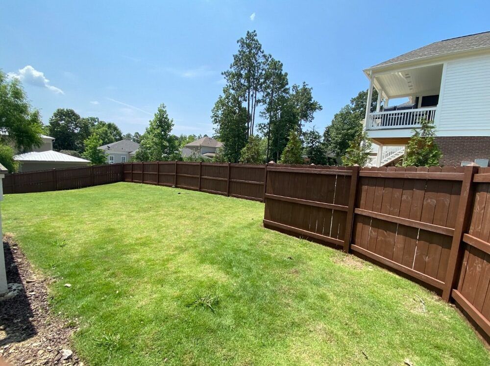 A brown wooden fence surrounds a green grassy yard.  A sunny day with trees and a house.