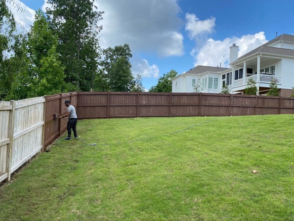 Person adjusting a brown wooden fence in a backyard with green grass and a white house under a cloudy sky.