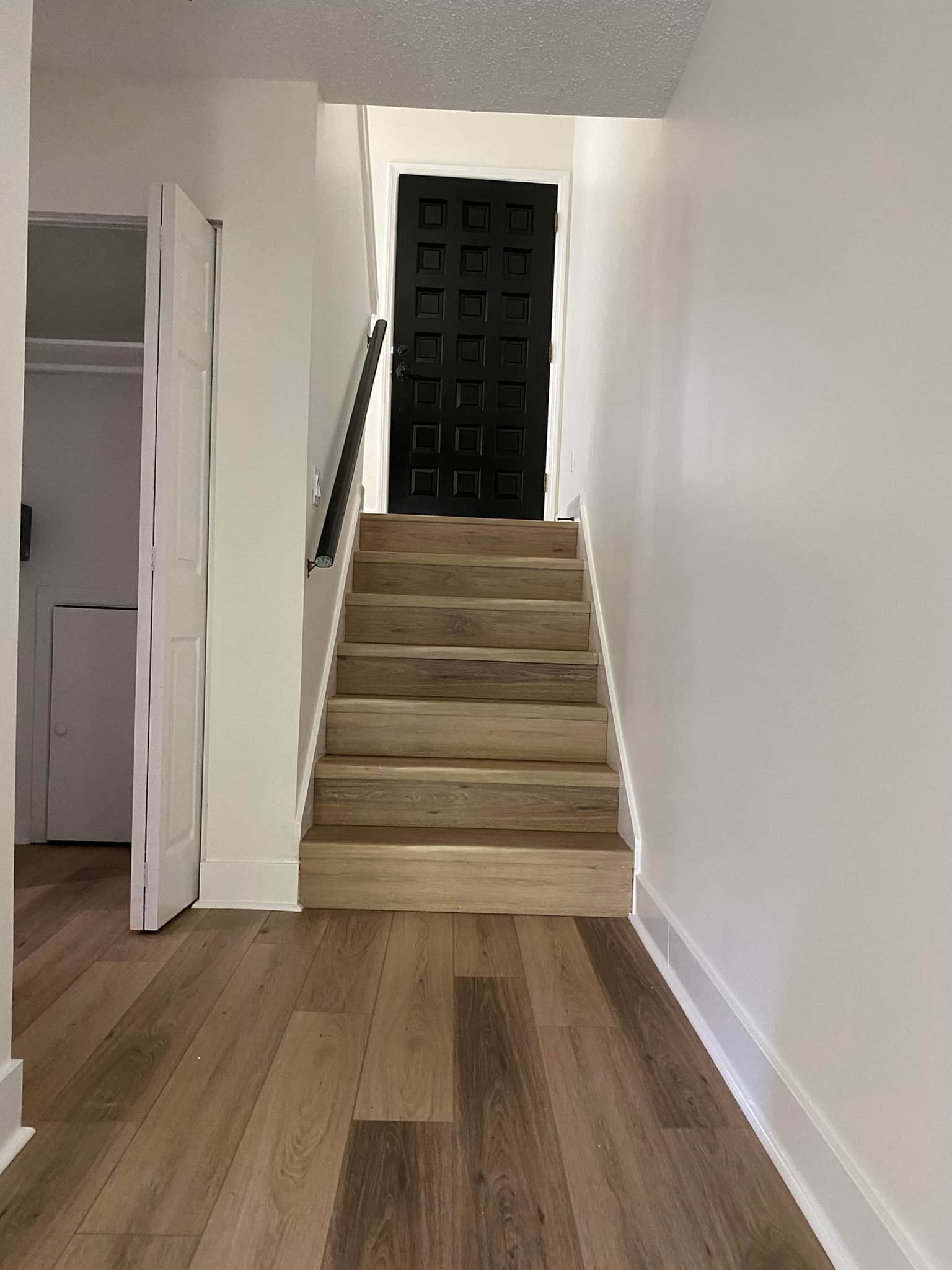 Interior view: wooden stairs leading up to a black door, with white walls and wood flooring.